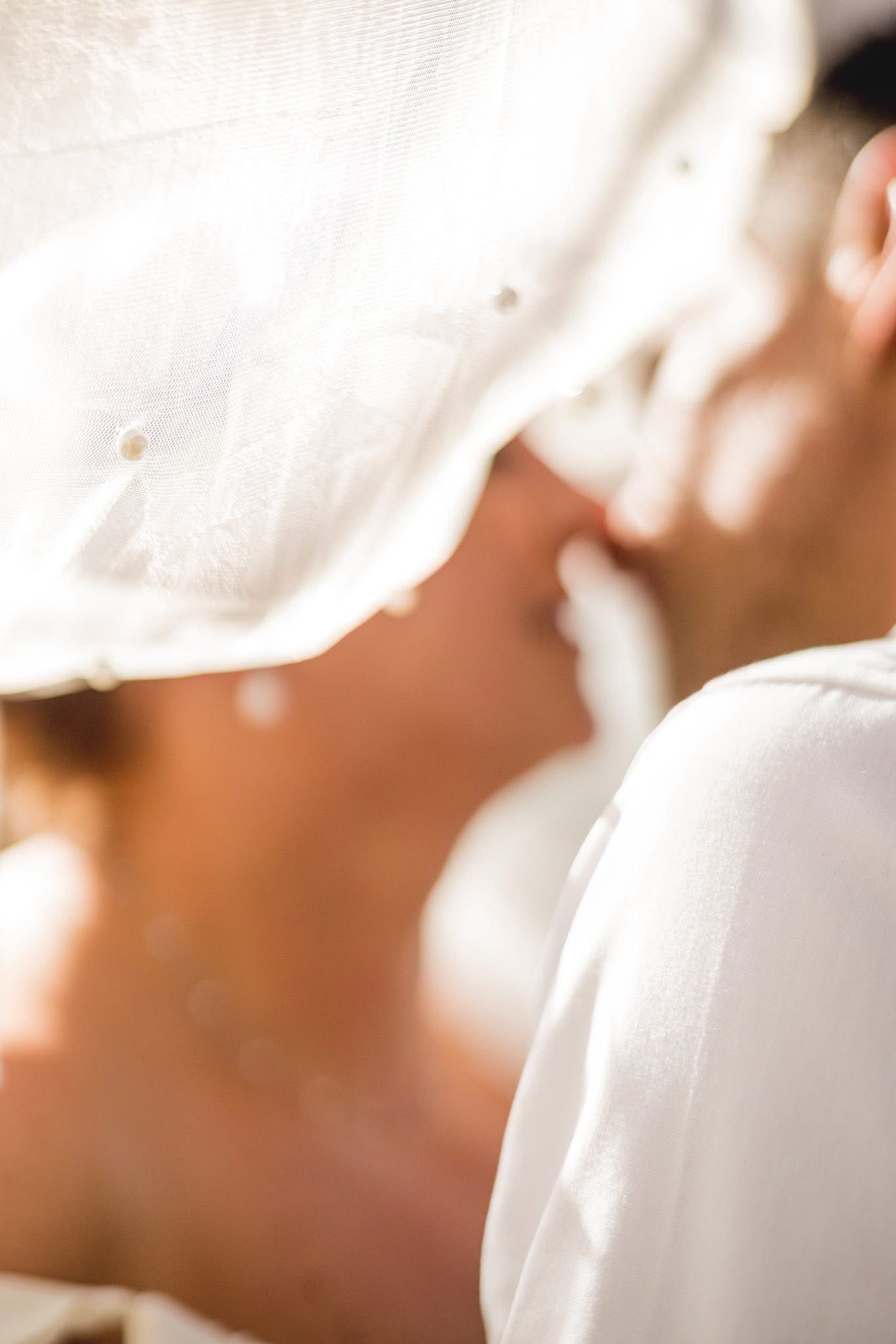 Romantic couple sharing a tender kiss under a white veil in soft sunlight.