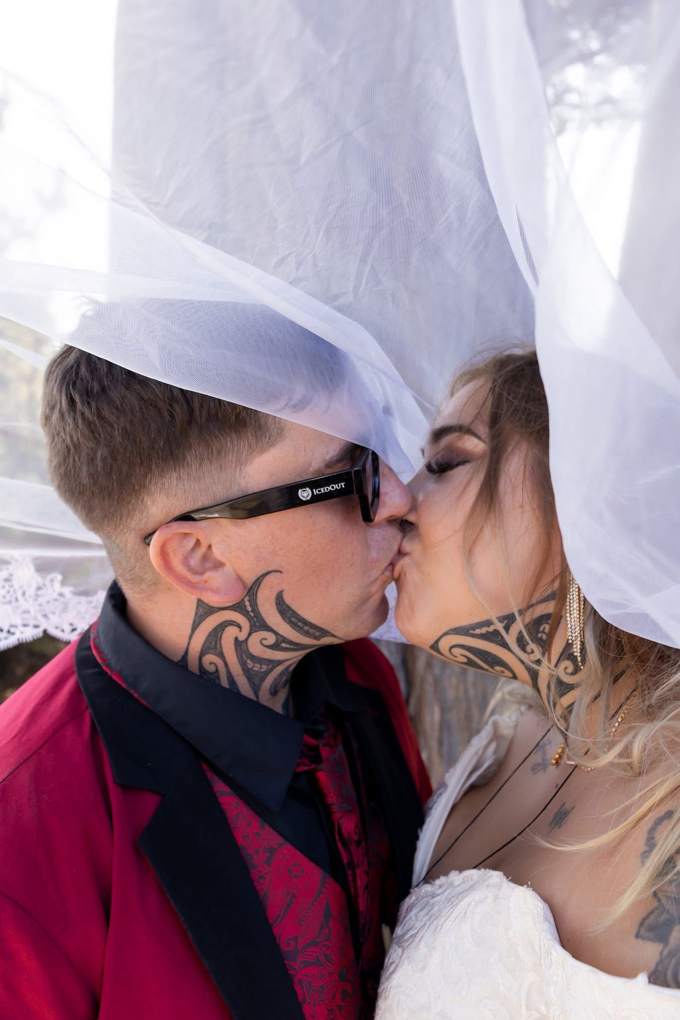 Tattooed bride and groom sharing a kiss under a white veil on their wedding day.