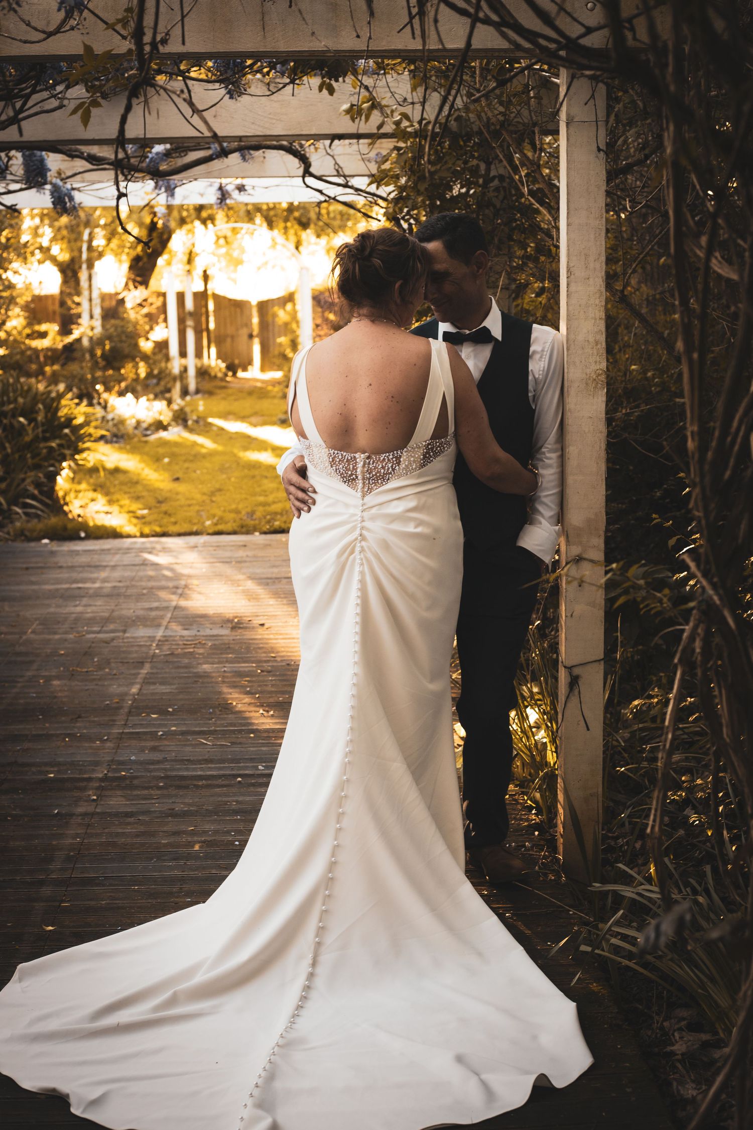 Bride and groom embrace under a wooden pergola in a lush garden, bride wearing an elegant backless white gown with a train.
