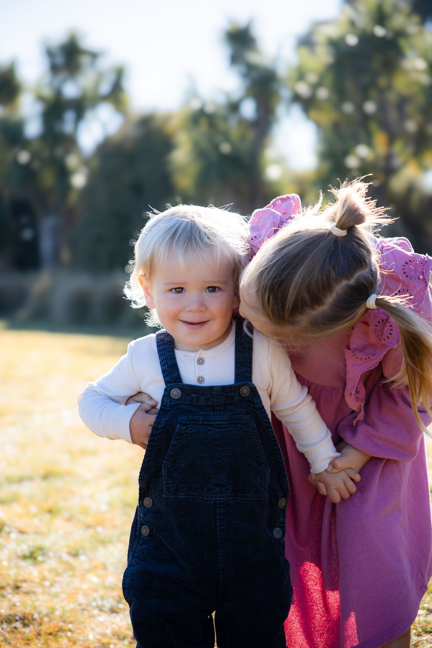 Two young siblings share a sweet moment outdoors, with an older girl in pink kissing her smiling toddler sibling in denim overalls.