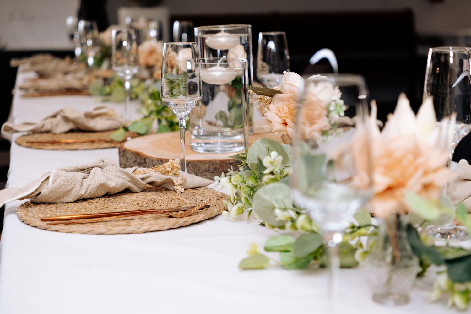 Elegant rustic wedding table setting with floral centerpieces, wood slice chargers, candles, and crystal glassware on white linen.