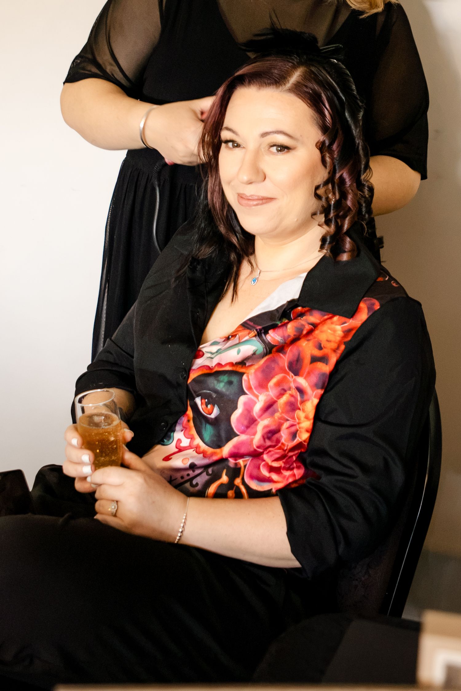 Bride relaxing with a drink while getting hair styled, wearing a colorful floral top before her wedding day.