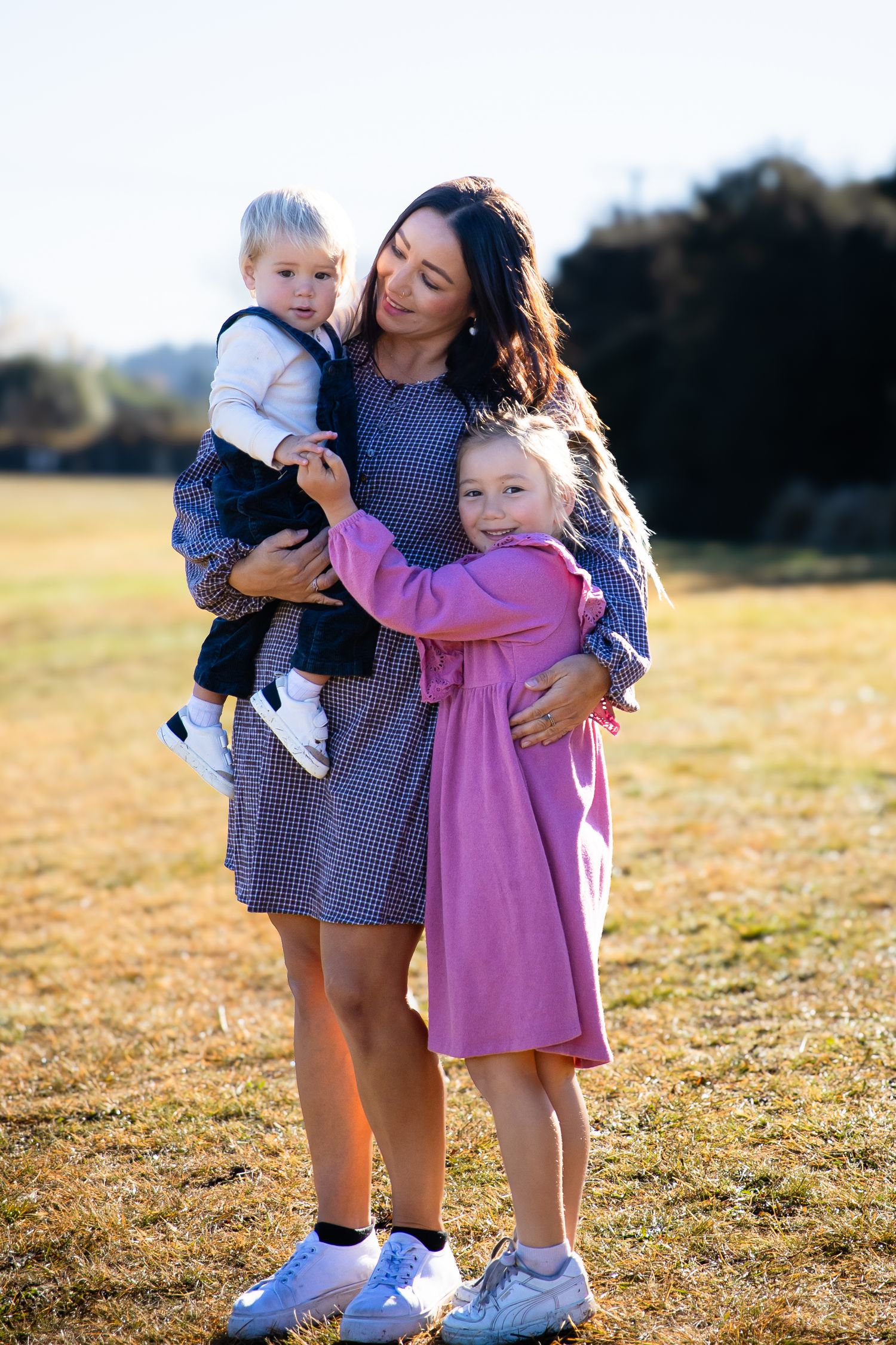 A mother in a blue dress holds her toddler son while her young daughter in pink hugs her in a sunny field.
