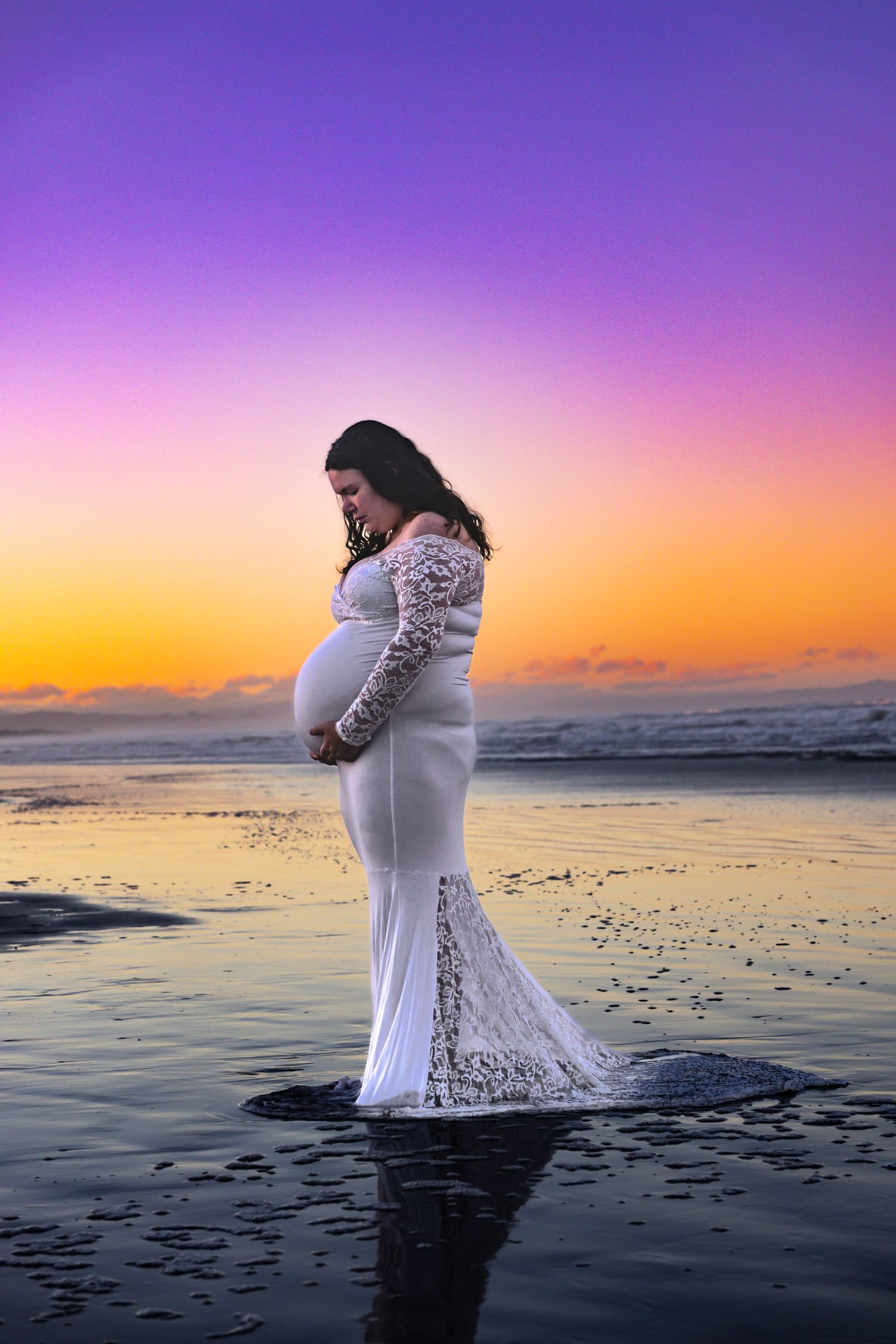 Pregnant woman in white lace gown standing on beach at sunset with purple and orange sky reflected in water.