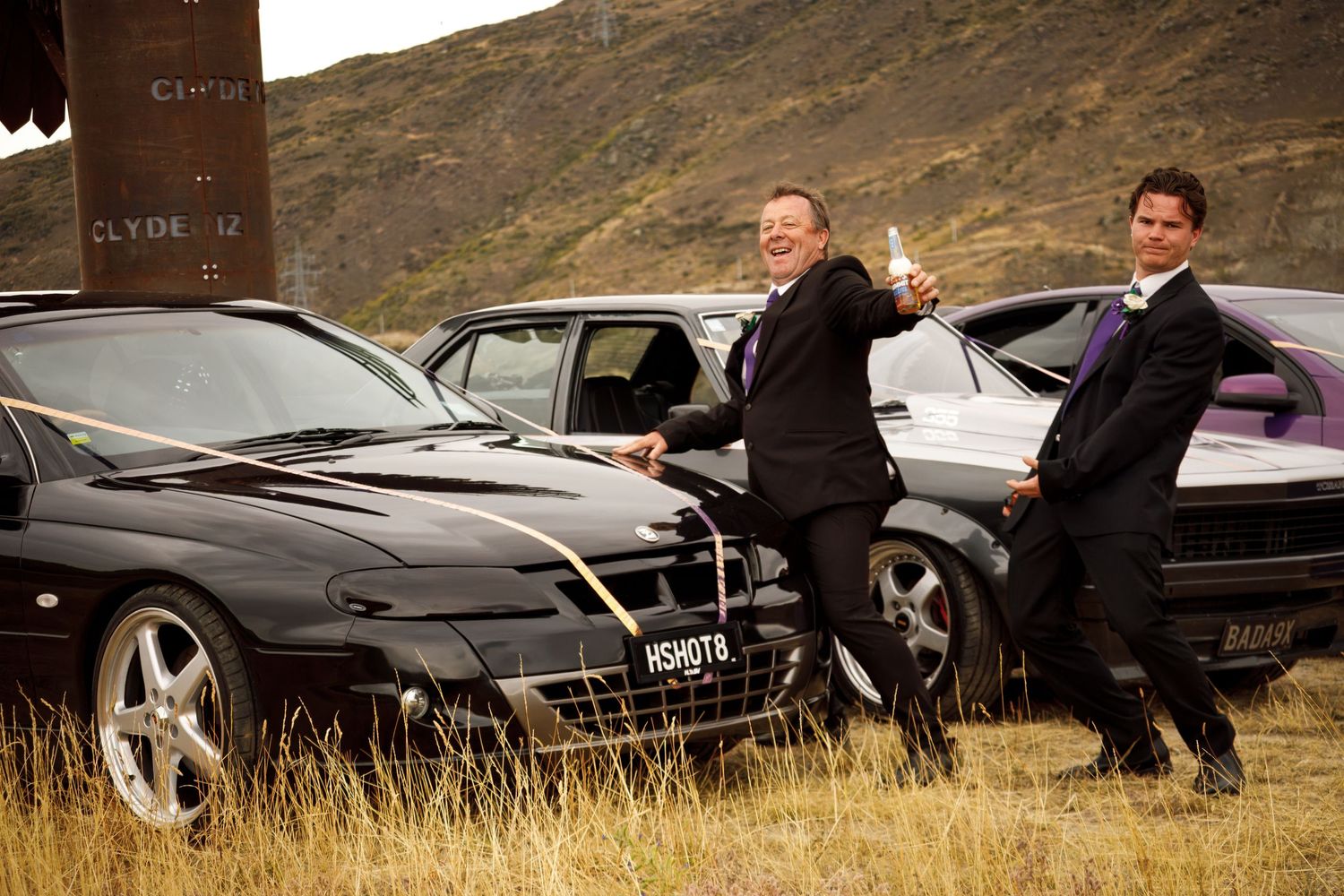 Two men in suits celebrate beside black luxury cars in a scenic mountainous landscape.