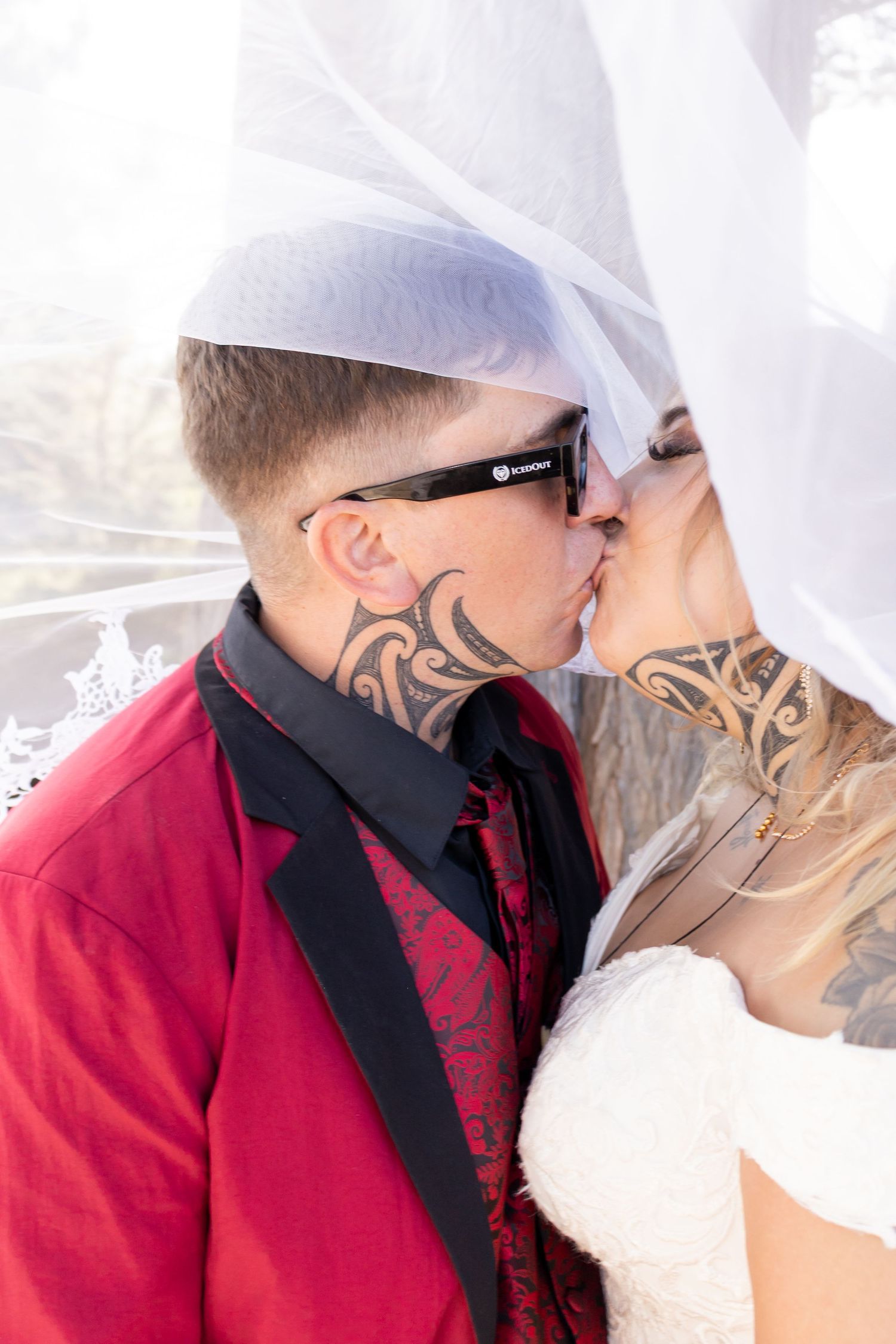 Tattooed bride and groom kissing under a white veil, groom in red suit with sunglasses at outdoor wedding.