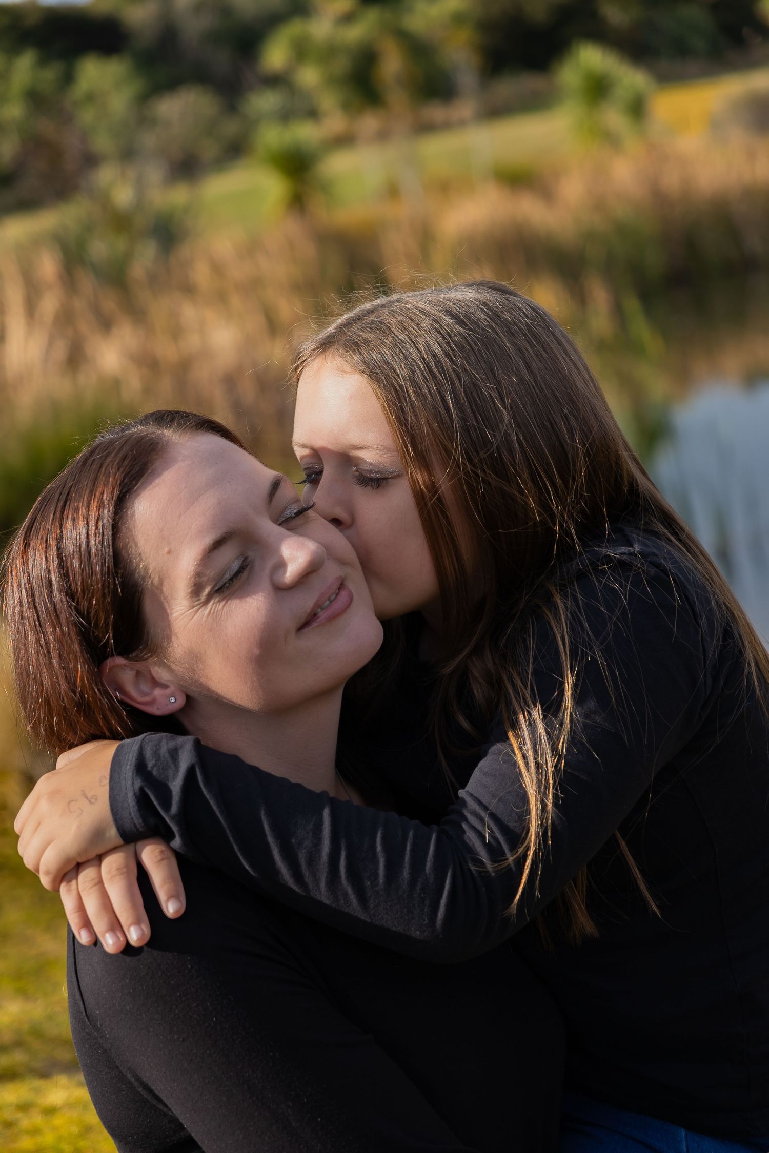 A young woman hugging and kissing an older woman on the cheek outdoors, both wearing black tops.
