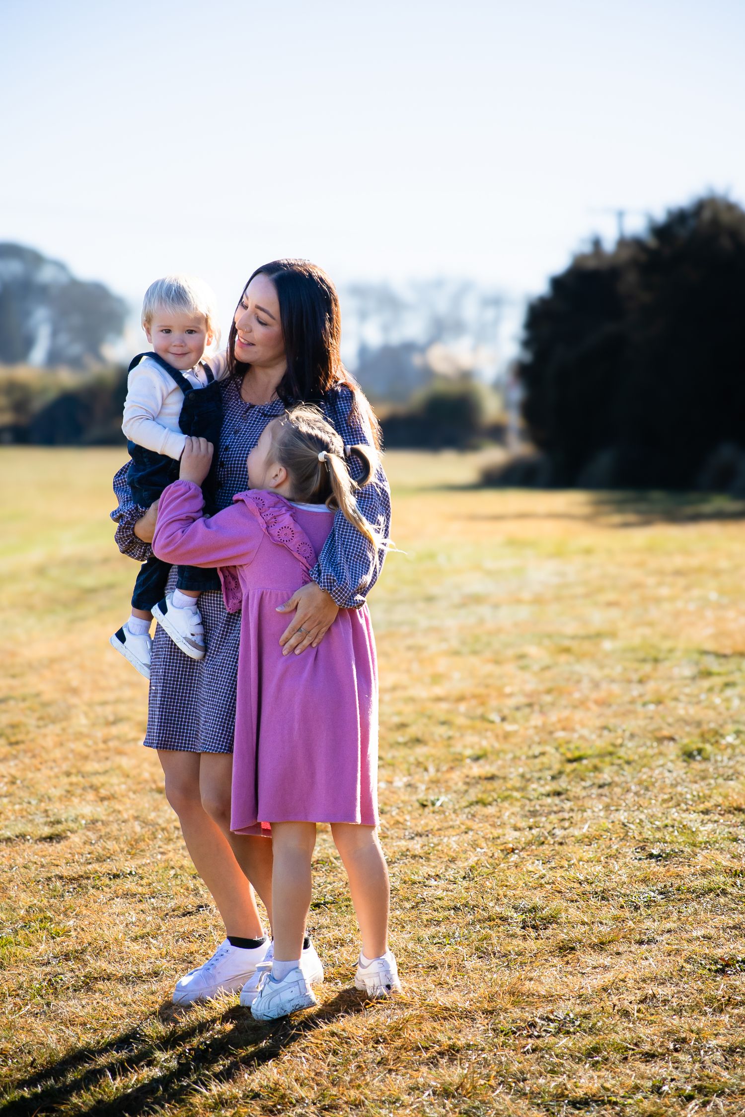 A mother holds her toddler son while her young daughter in a pink dress hugs her in a sunny green park.