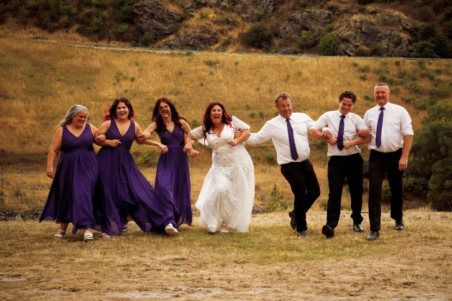 Wedding party in purple bridesmaid dresses and suits running joyfully across a golden field on a sunny day.