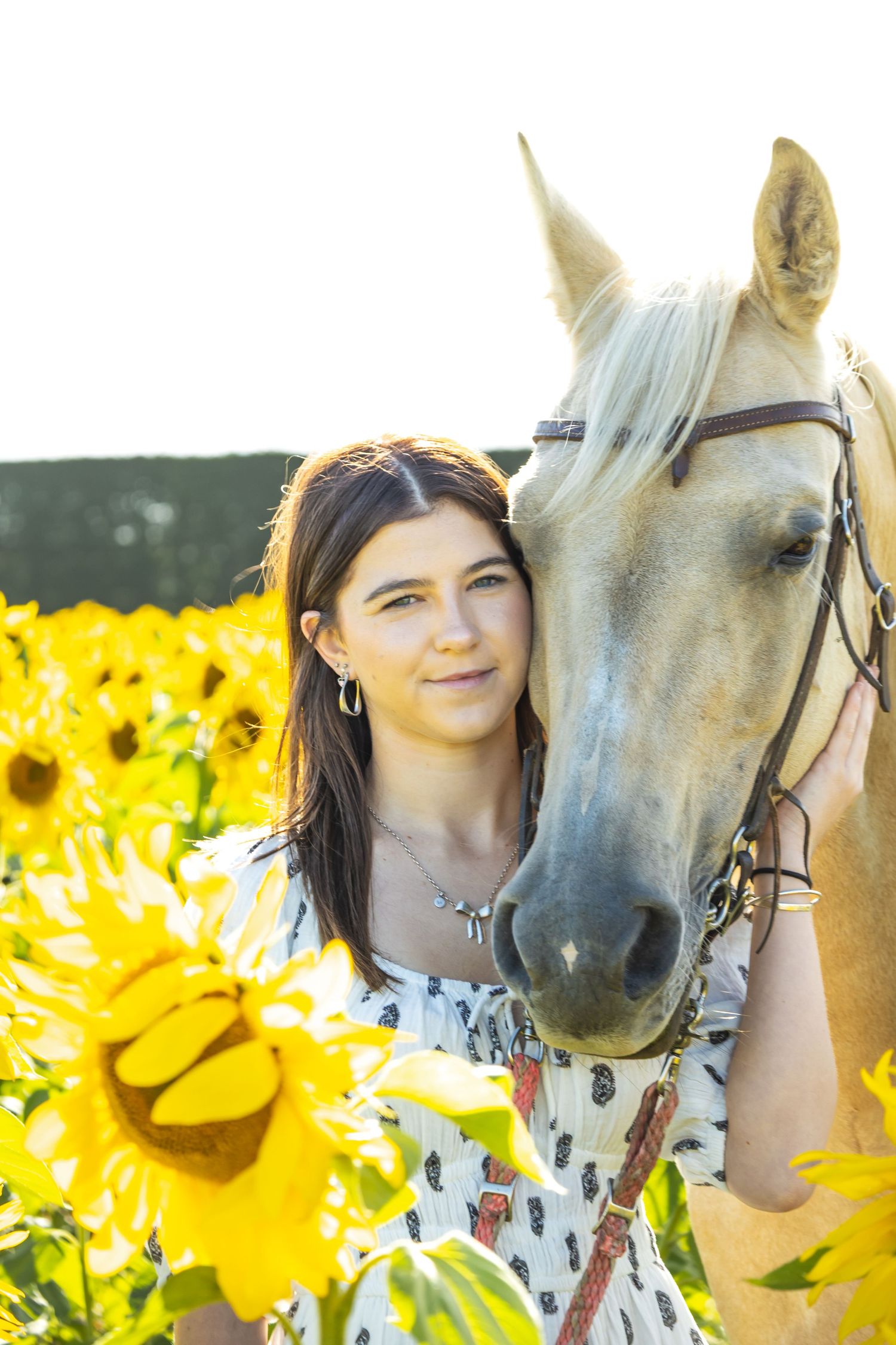 Young woman in floral dress poses with palomino horse in vibrant sunflower field on bright sunny day.