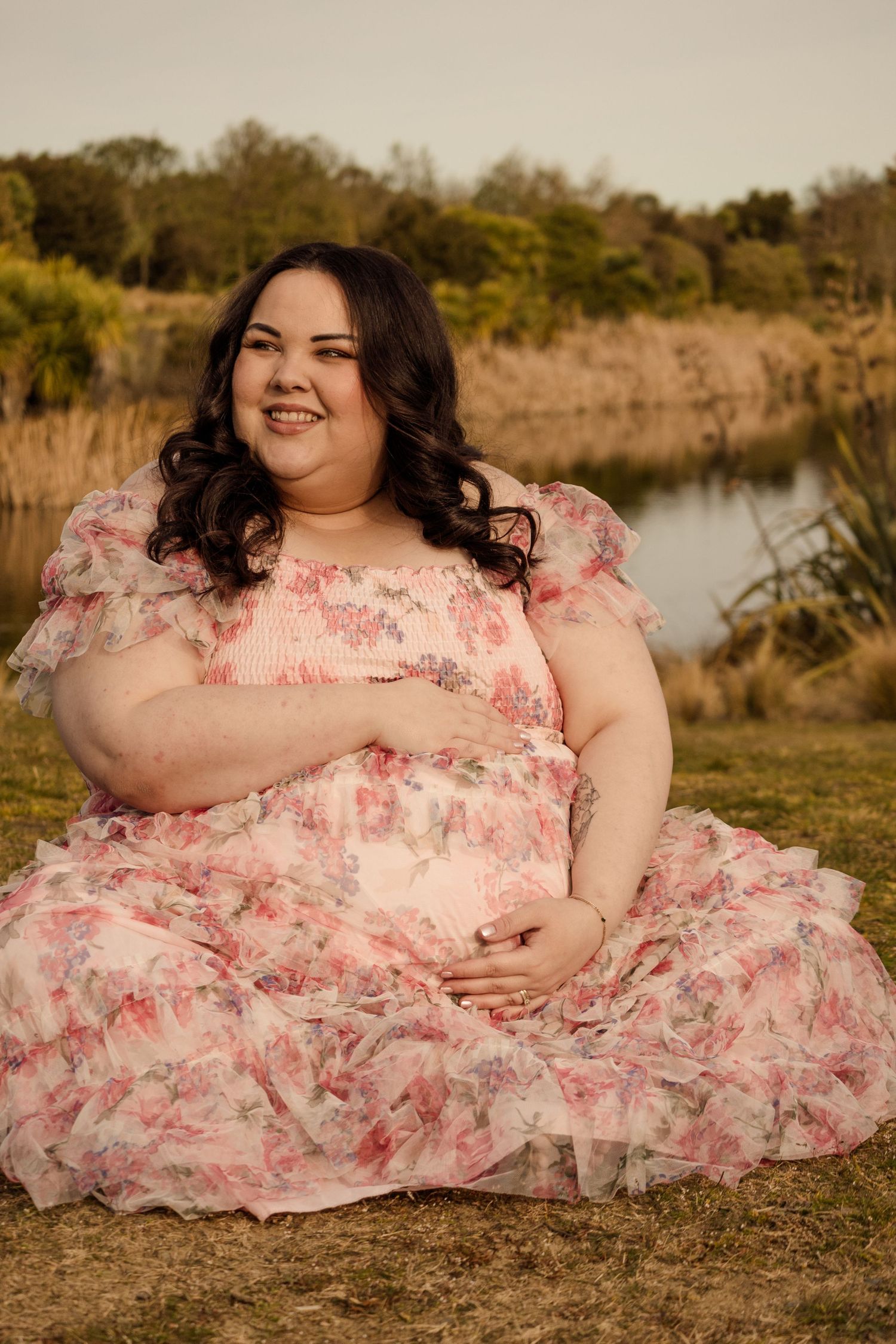 Plus-size woman in floral pink gown sitting outdoors by a lake, smiling joyfully in a lush green setting.