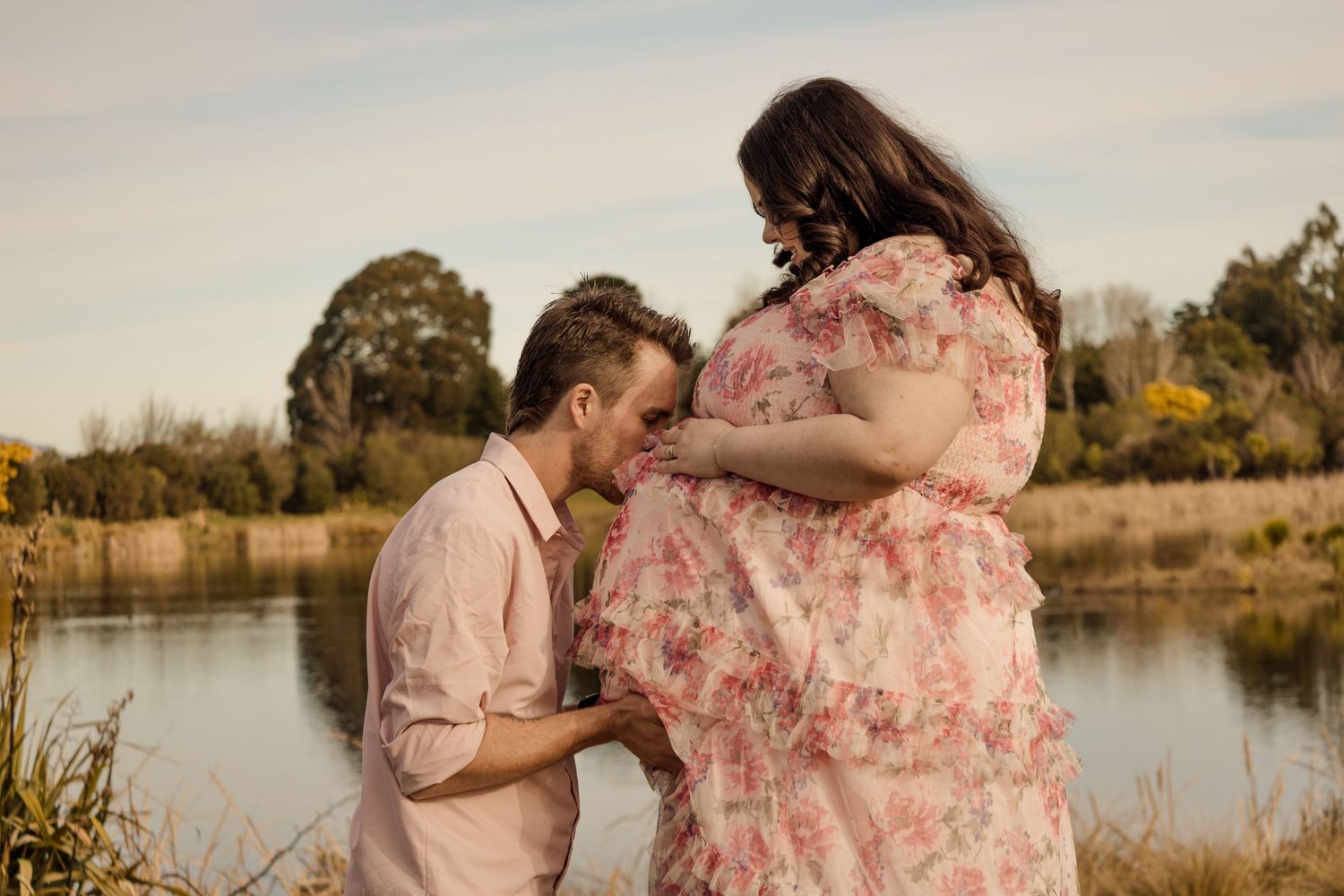 A man kisses a pregnant woman's belly outdoors near a serene lake, both dressed in soft pink attire.
