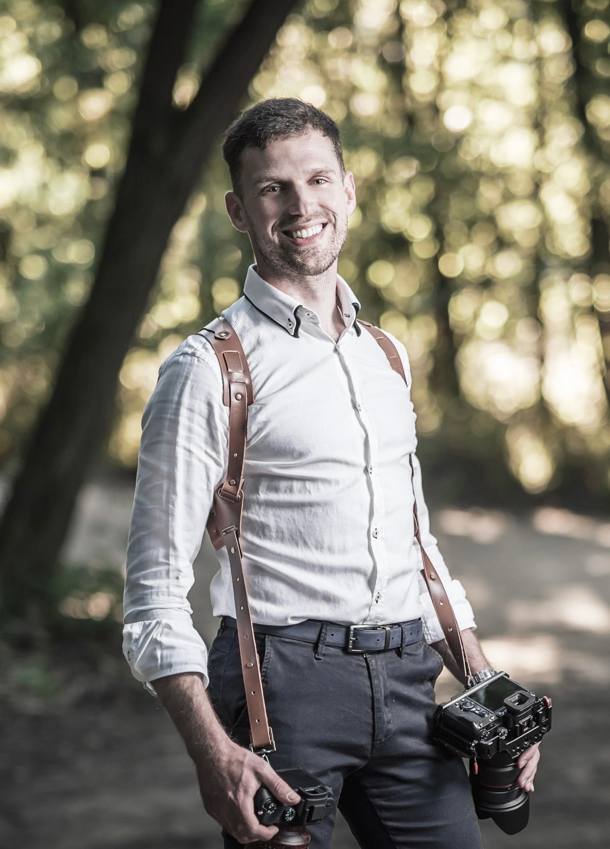 Wedding photographer in white shirt and suspenders holding cameras in wooded area.