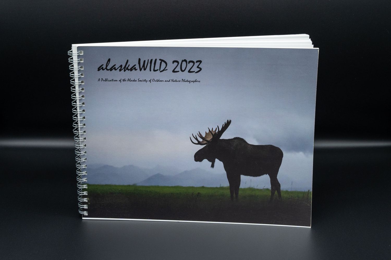 A photograph of the alaskaWILD 2023 booklet. The booklet cover shows the Best in Show image of a moose in silhouette.