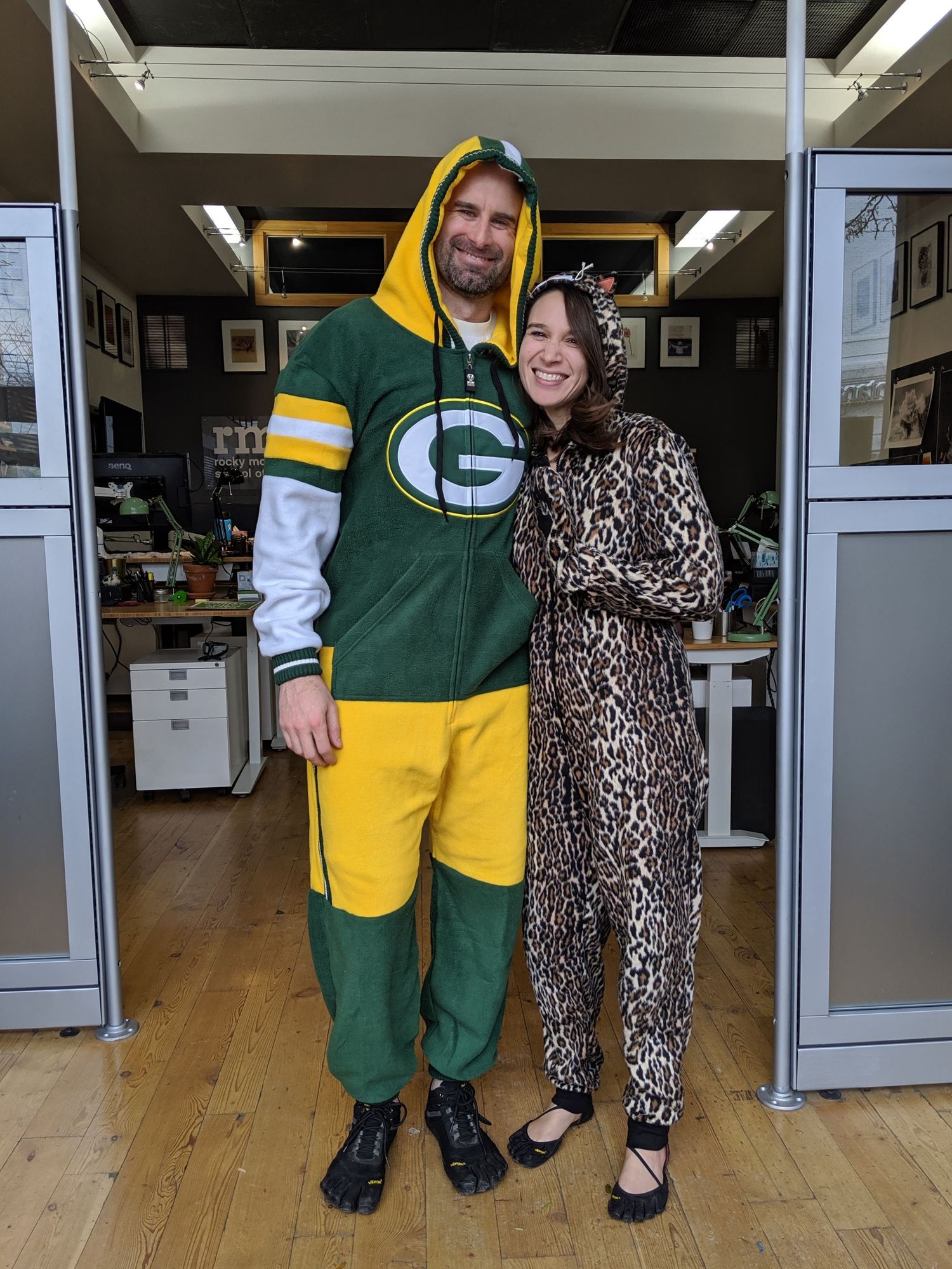 A man and woman in onesies. The man is in a Green Bay Packers onesie. The woman is in a cat onesie.