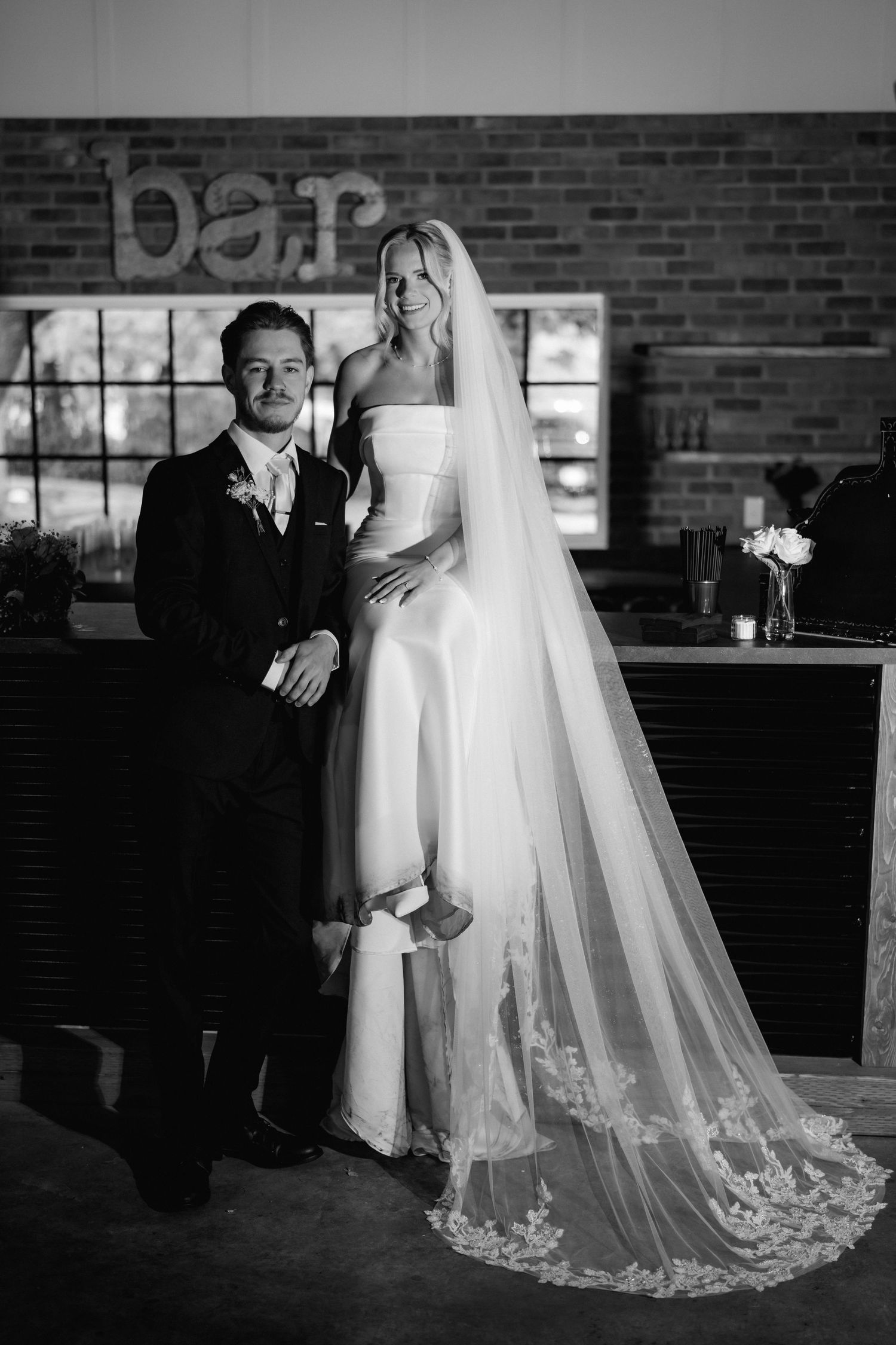 Black and white wedding photo of bride in strapless gown with lace veil and groom in suit posing by a bar.