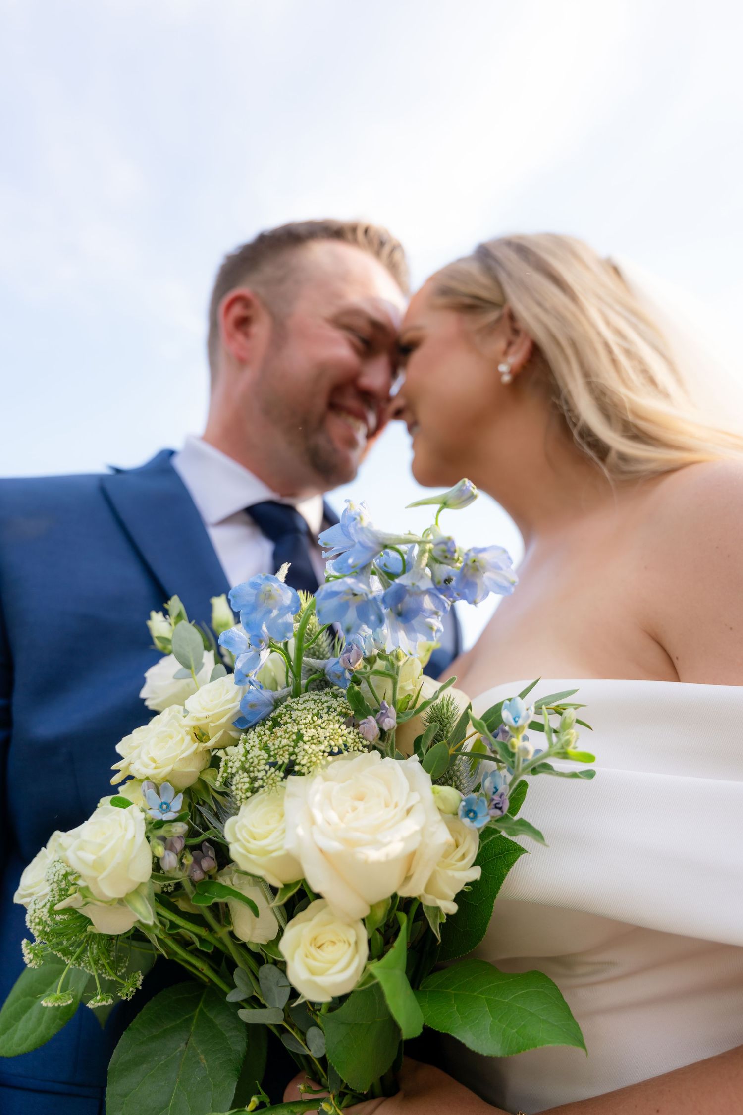 Bride and groom touching foreheads smiling with blue and white wedding bouquet in foreground.