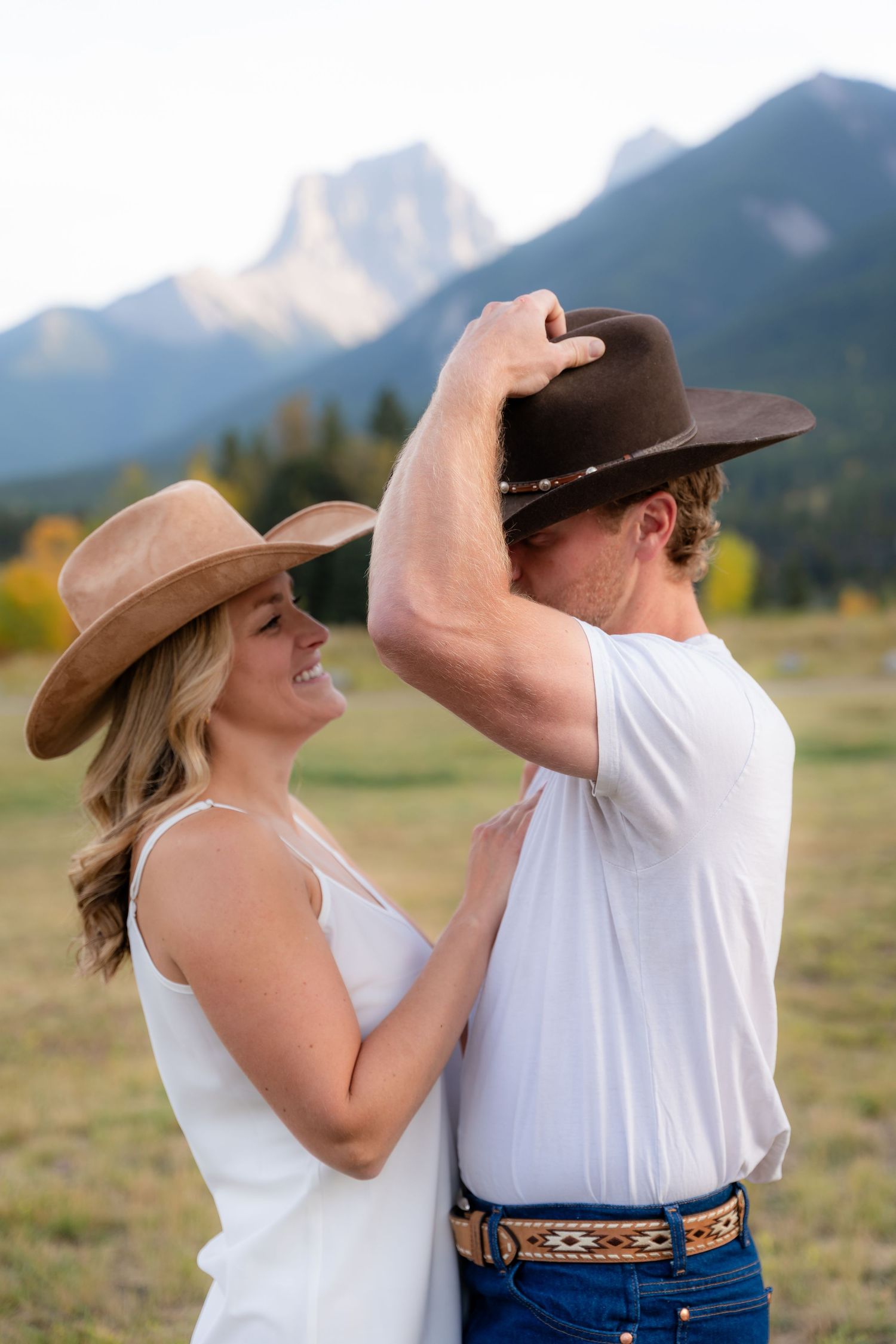 Couple in cowboy hats embracing in a mountain meadow, woman smiling in white dress, man in white tee and jeans.