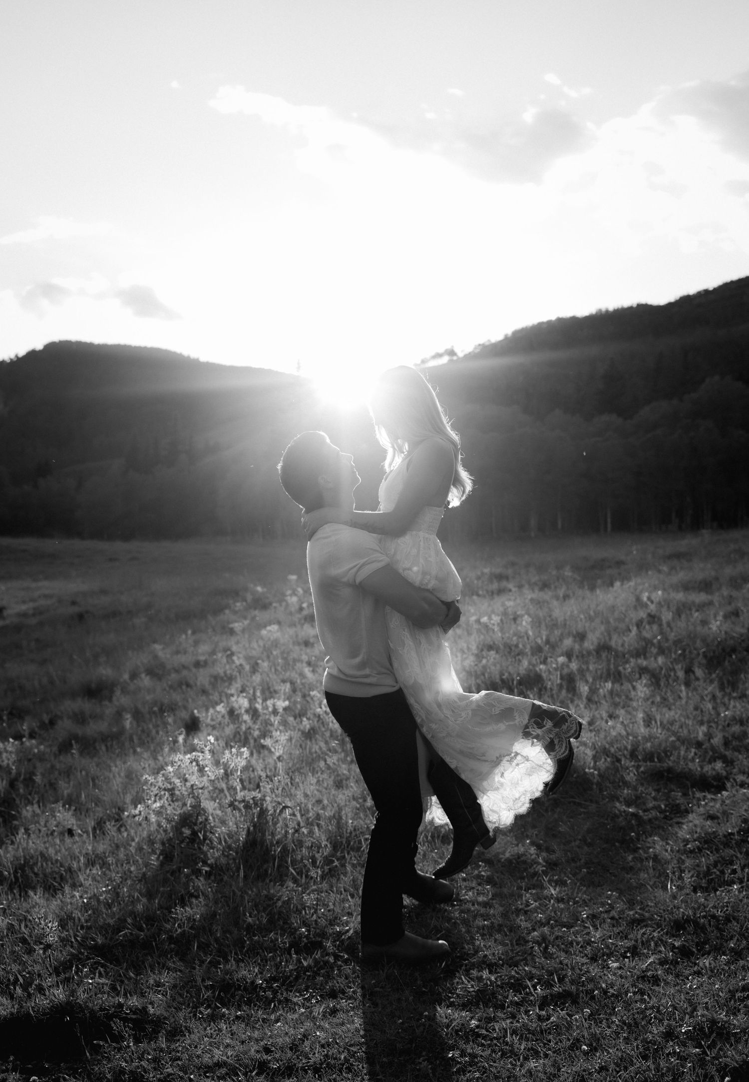 Couple embracing in a sunlit field with mountains in the background in black and white.