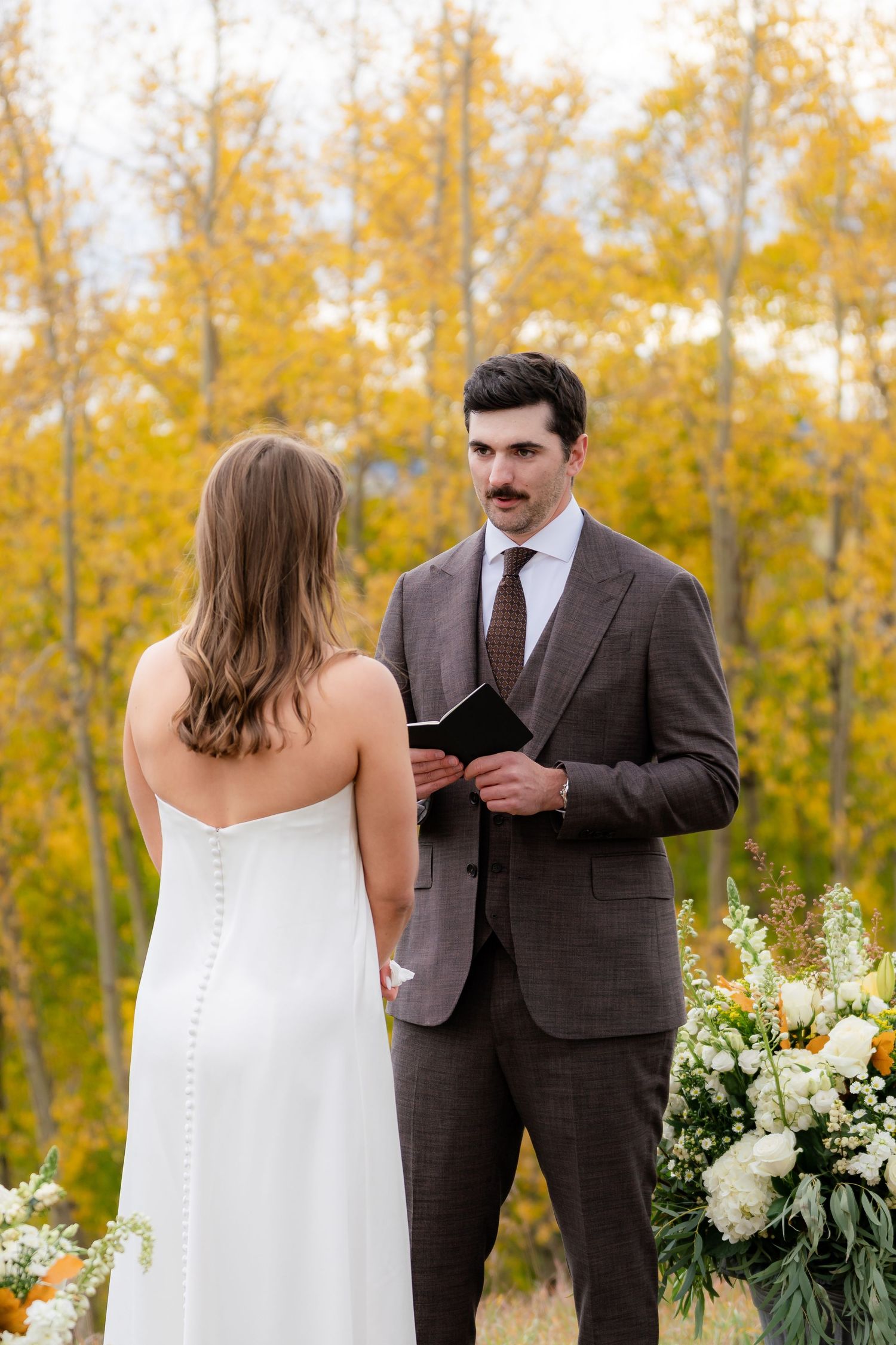 Groom with mustache reading vows to bride outdoors among golden autumn trees.