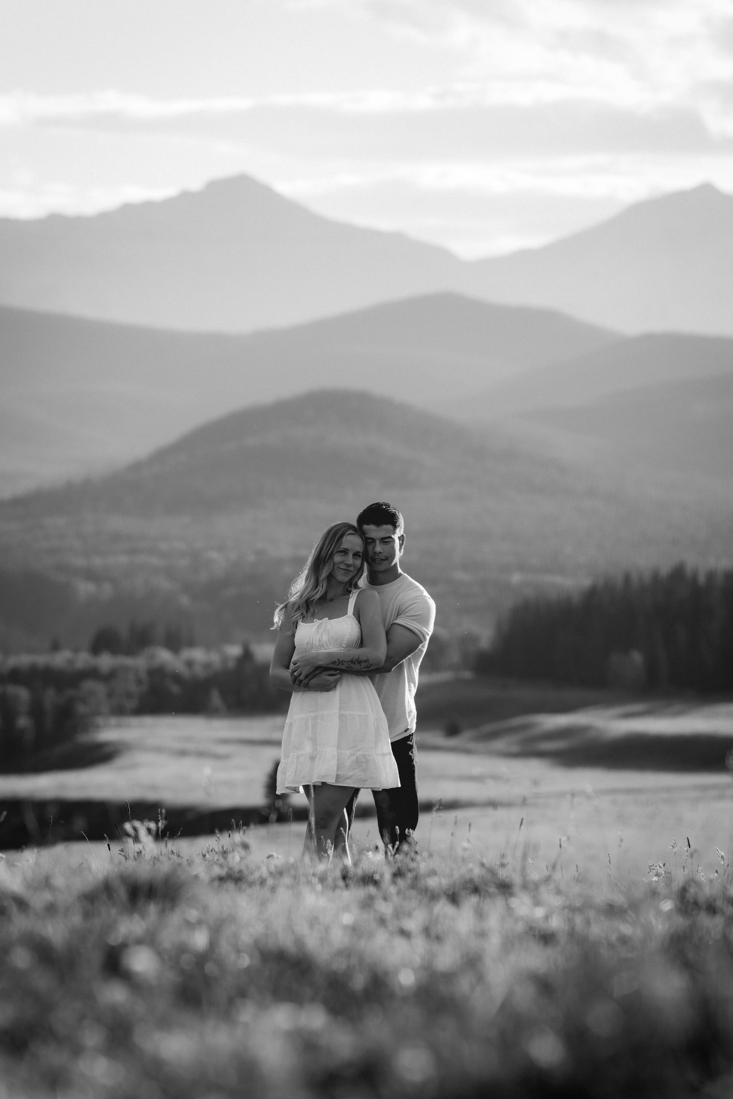 Black and white image of a couple embracing in a meadow with dramatic layered mountain peaks in the background.