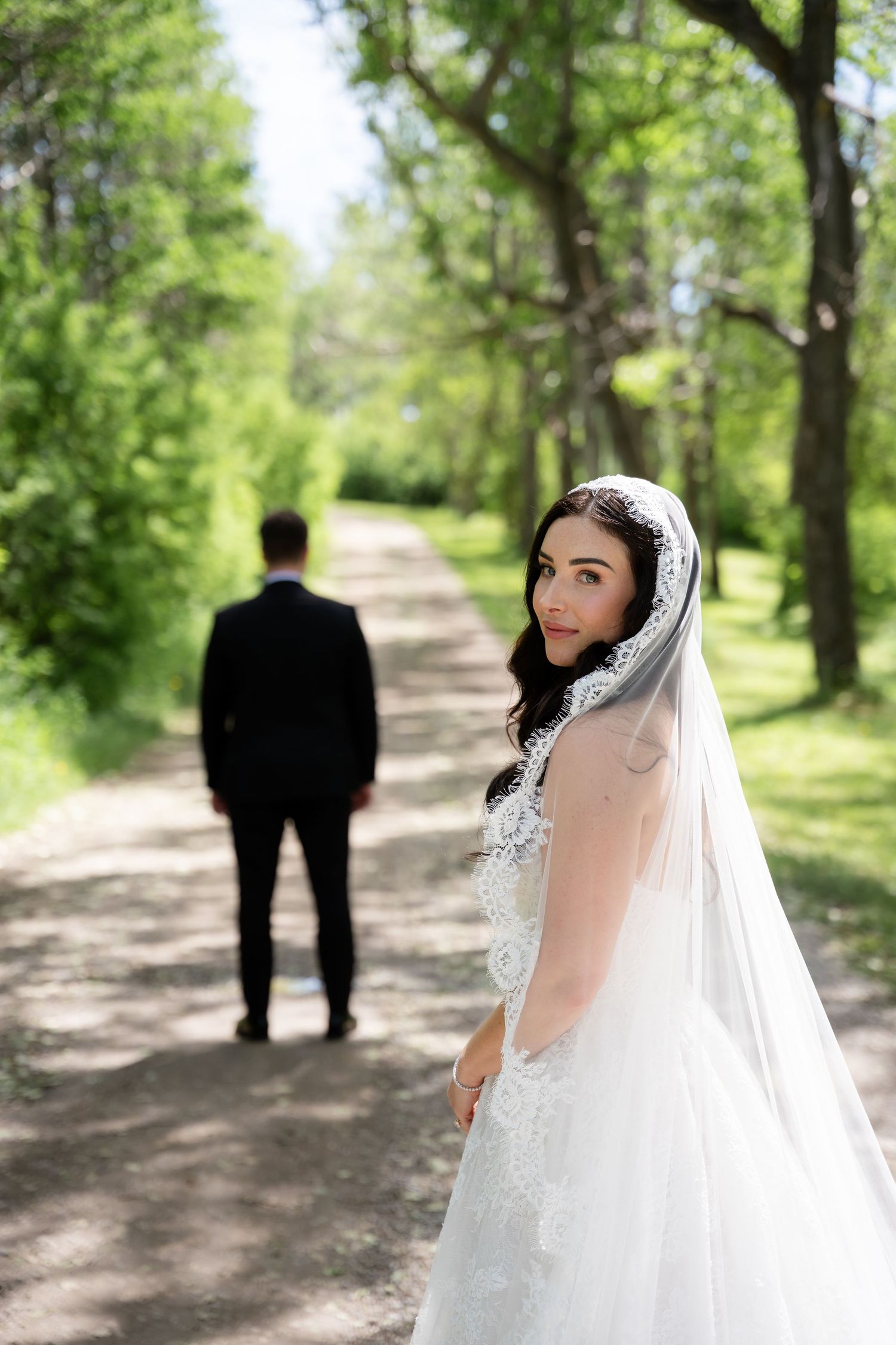Bride in white lace gown and veil looks back while groom stands on tree-lined path on wedding day.