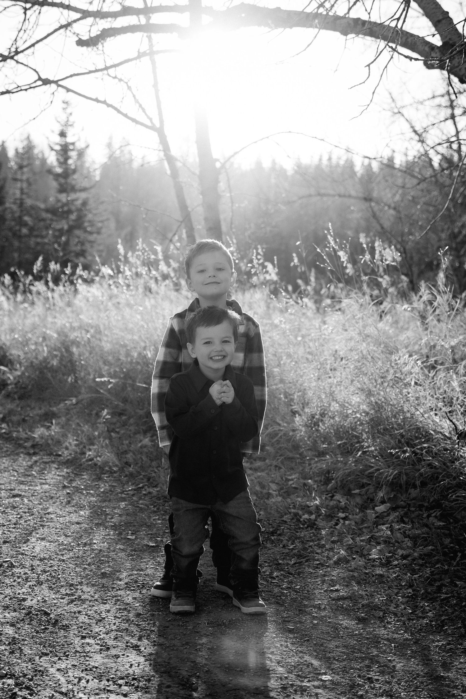 Two young boys smiling on a woodland path, black and white portrait with sunlight filtering through trees.