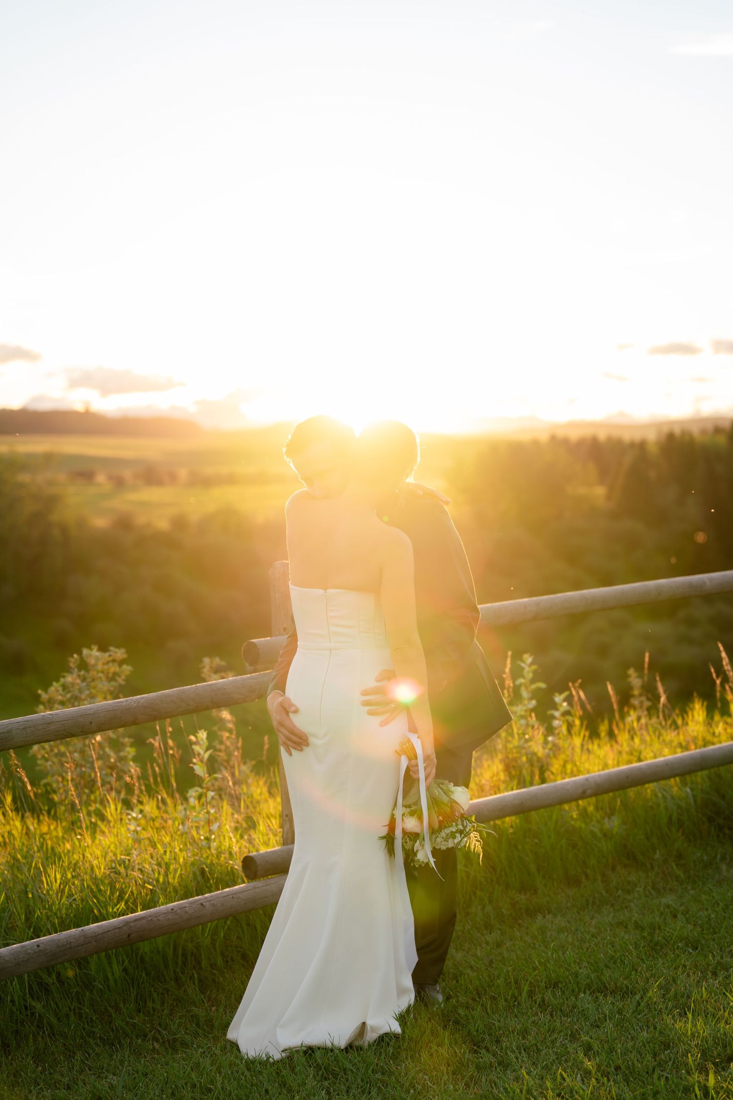 Bride and groom silhouetted against golden sunset overlooking lush green countryside landscape.