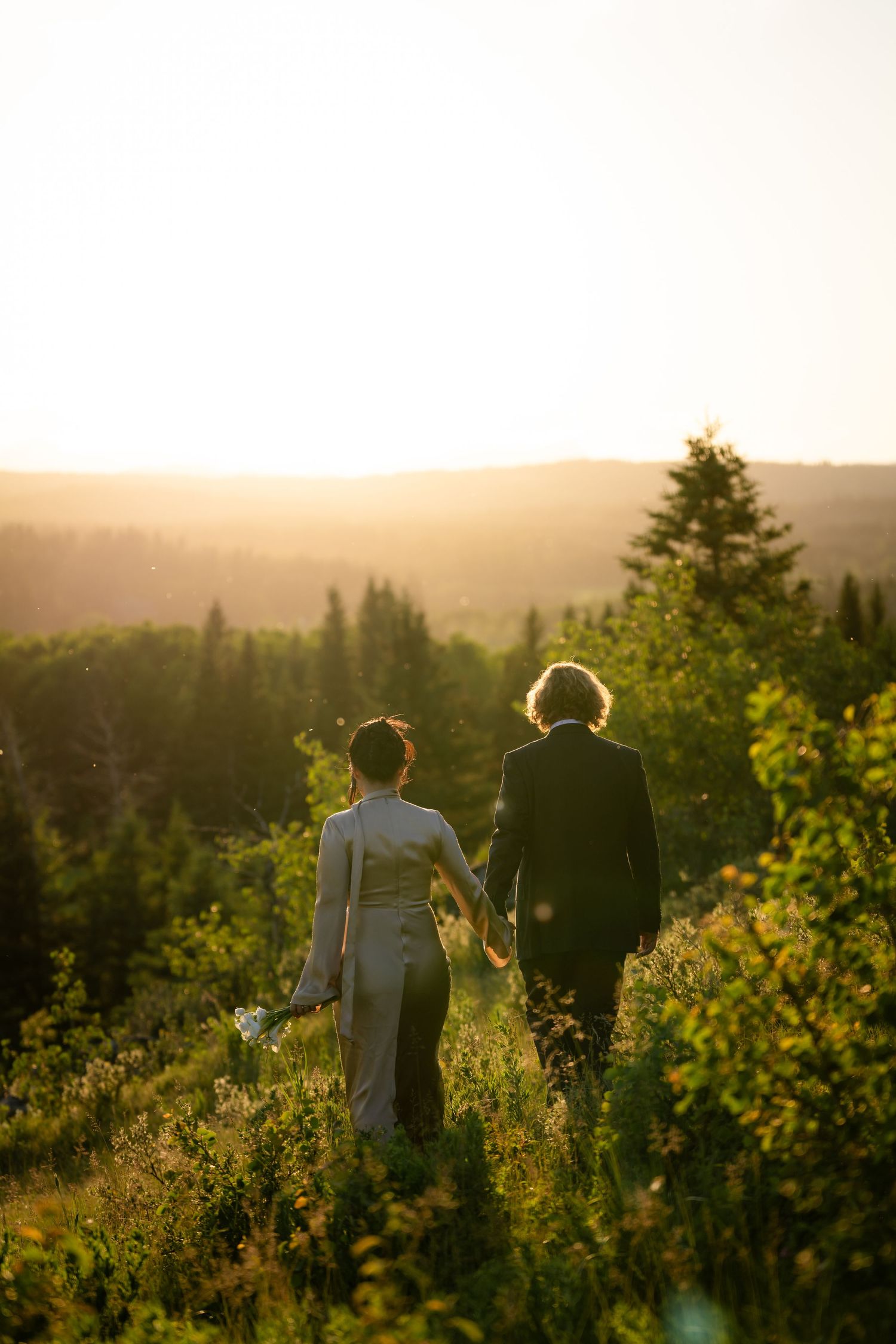 Couple holding hands walking through lush green meadow at golden sunset with forest backdrop.