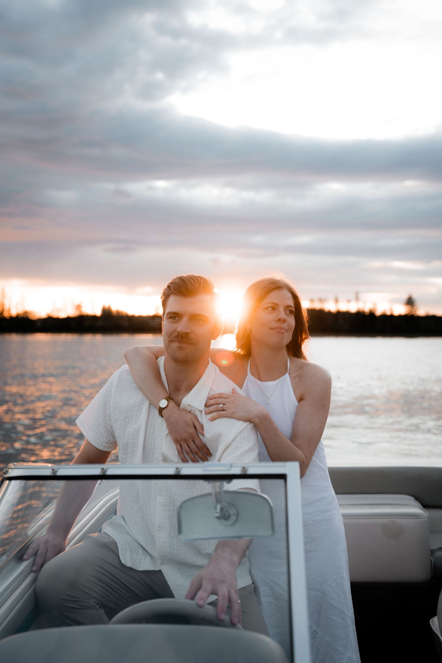 Couple in white outfits on a boat at sunset, golden light reflecting on calm water behind them.