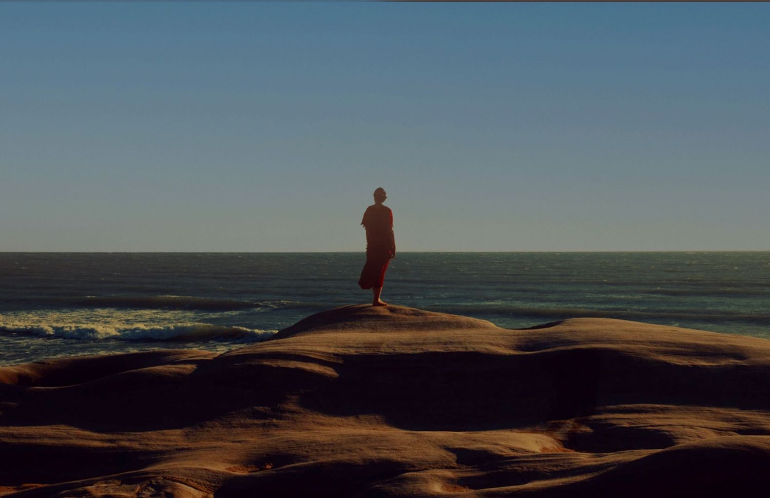 A lone figure stands on rocky terrain overlooking a vast ocean under a clear blue sky.
