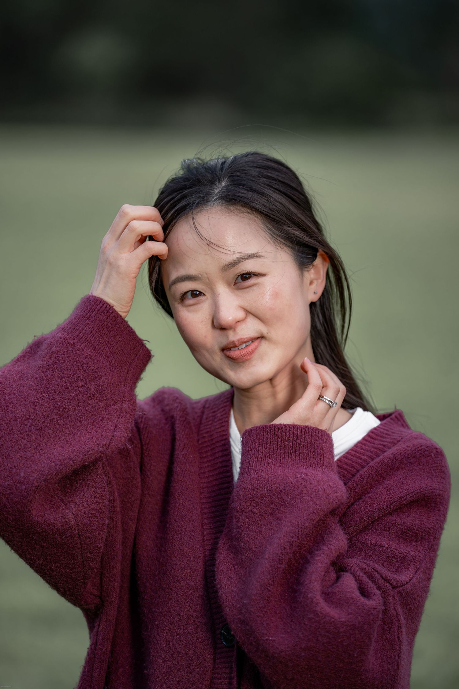 A young Asian woman in a burgundy sweater poses outdoors, touching her hair and neck, smiling softly.