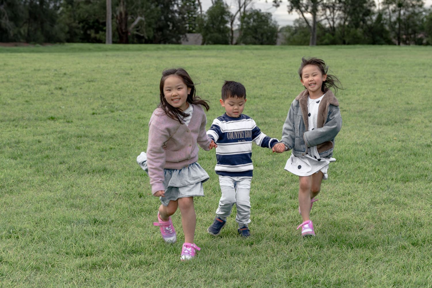 Three happy Asian children running and holding hands on a lush green grass field outdoors.