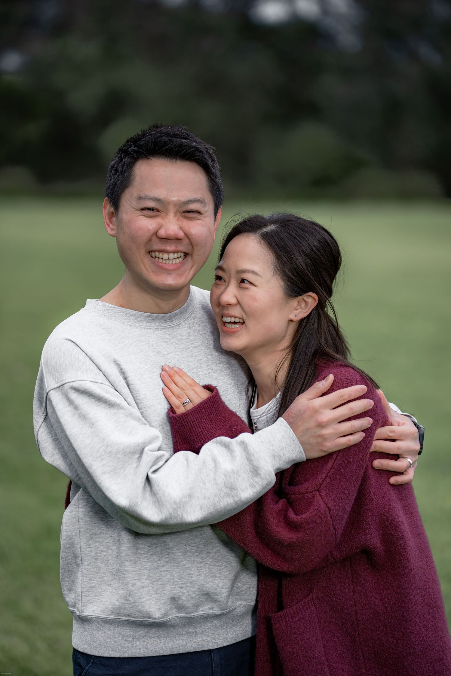 Happy couple embracing outdoors in a park, smiling joyfully, woman wearing a ring on her finger.