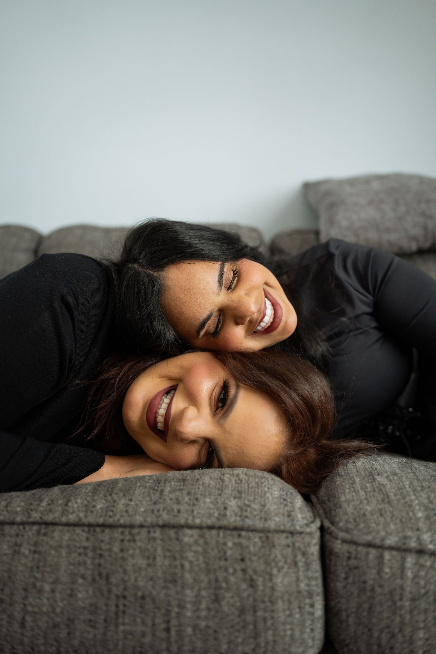 Two people in black tops share joyful laughter while relaxing together on a gray couch.