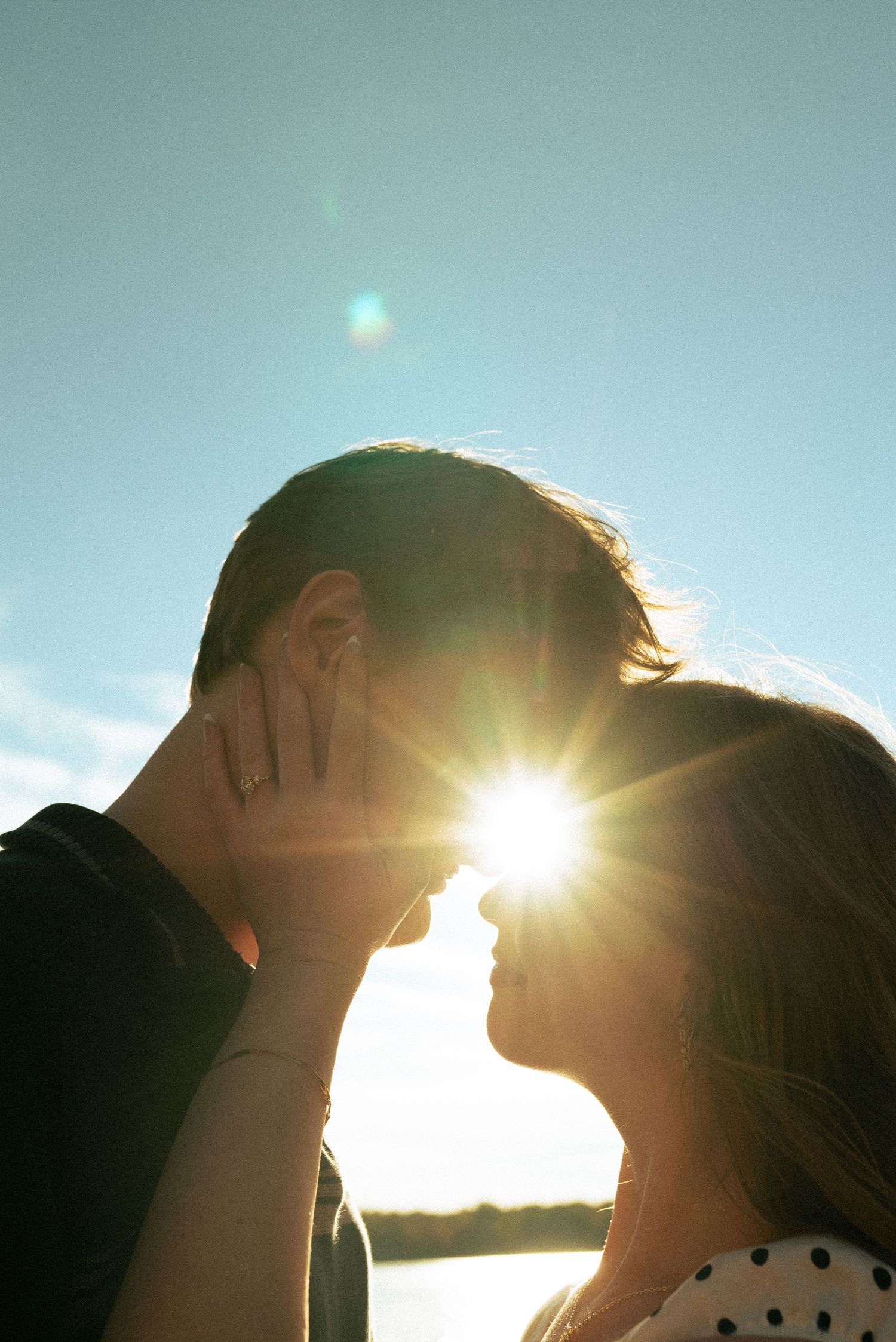 Engaged couple with sun flare behind them. Hand with ring on mans face.