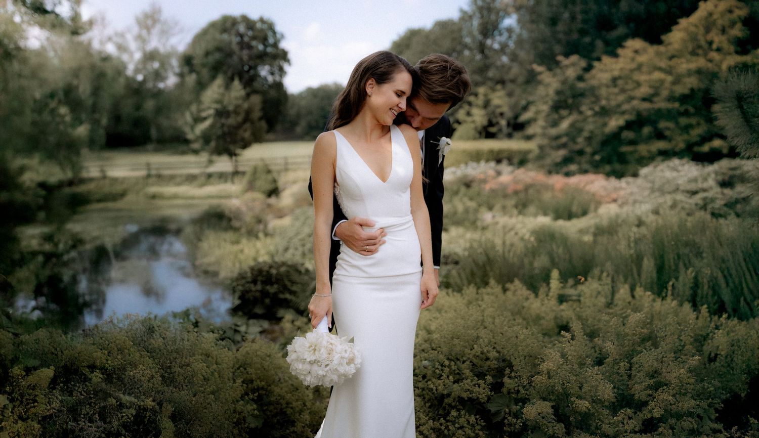A romantic couple embraces by a serene pond surrounded by lush greenery on their wedding day.