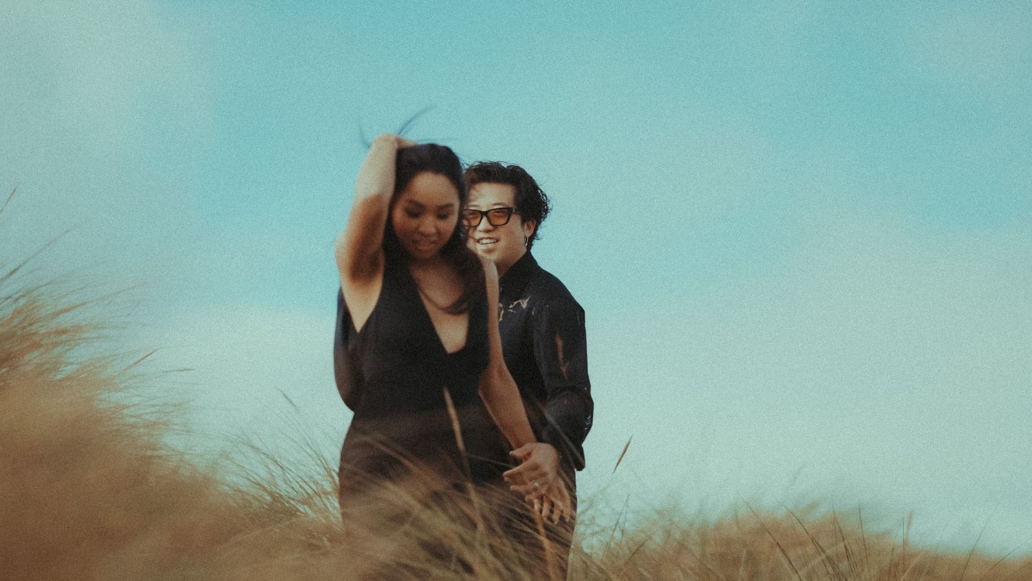 A couple stands together in tall grass against a turquoise sky during golden hour.