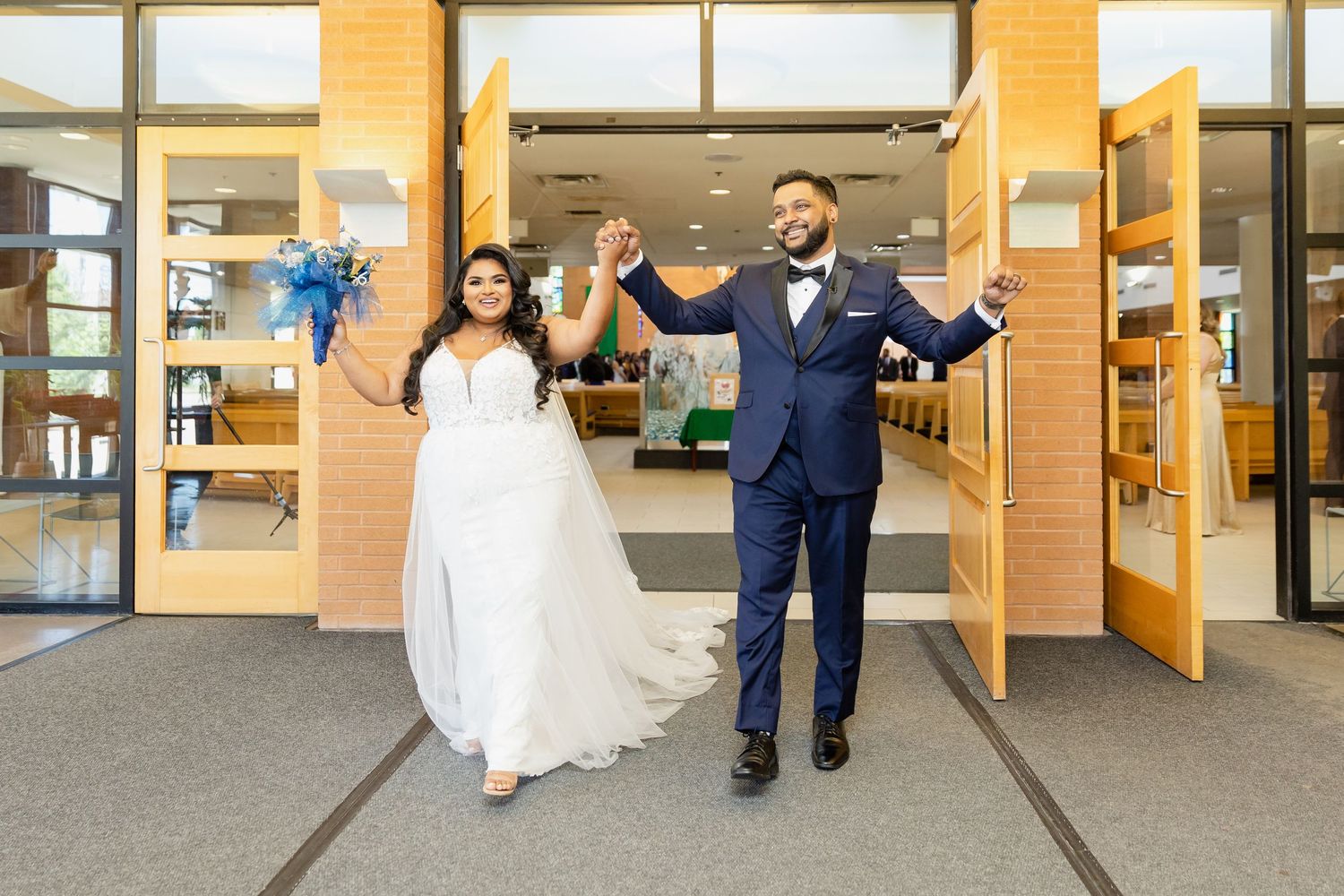 A couple exits through doors in wedding attire with joyful expressions.