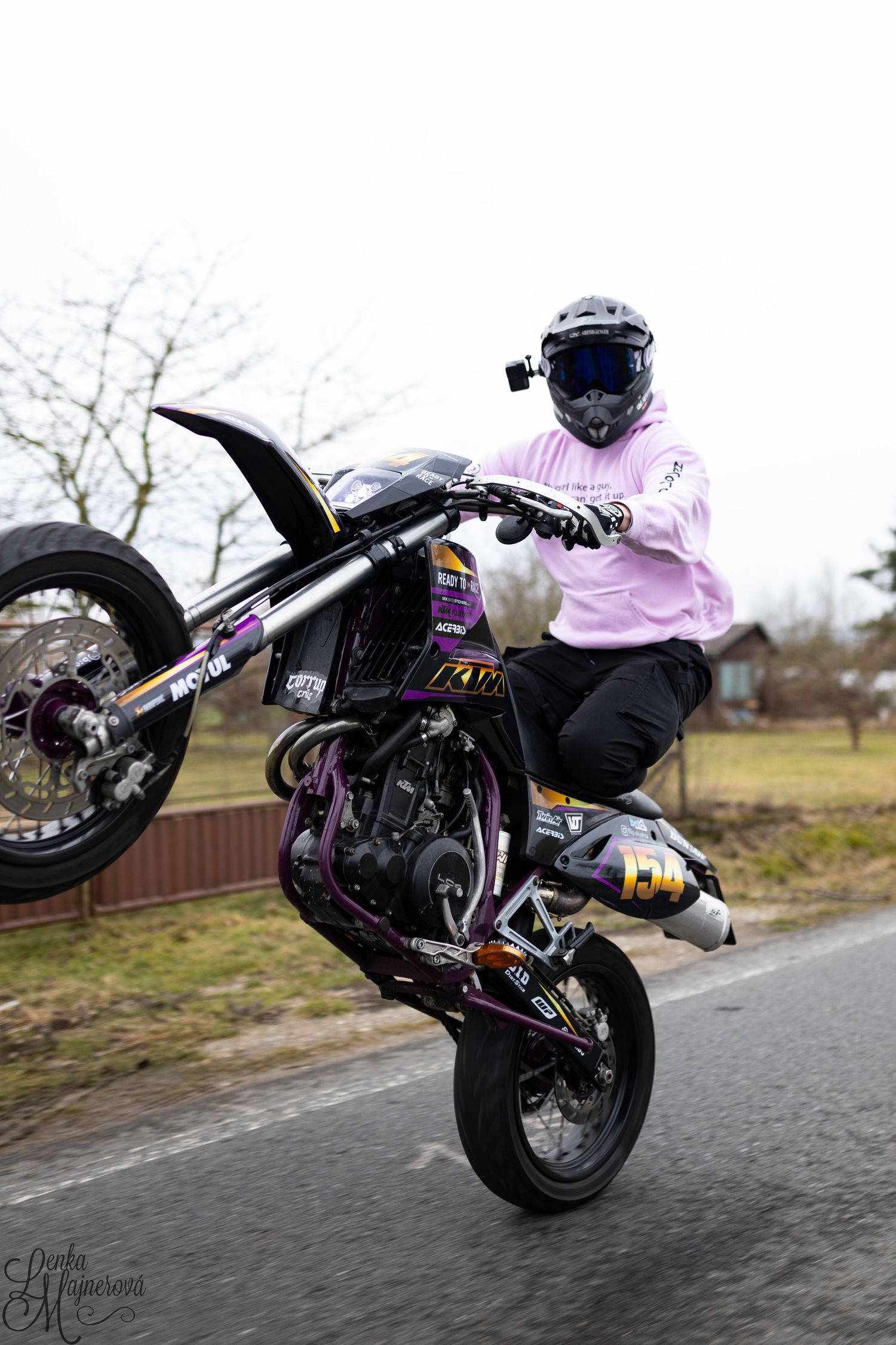 Stunt rider performing wheelie on black motorcycle wearing pink sweatshirt.