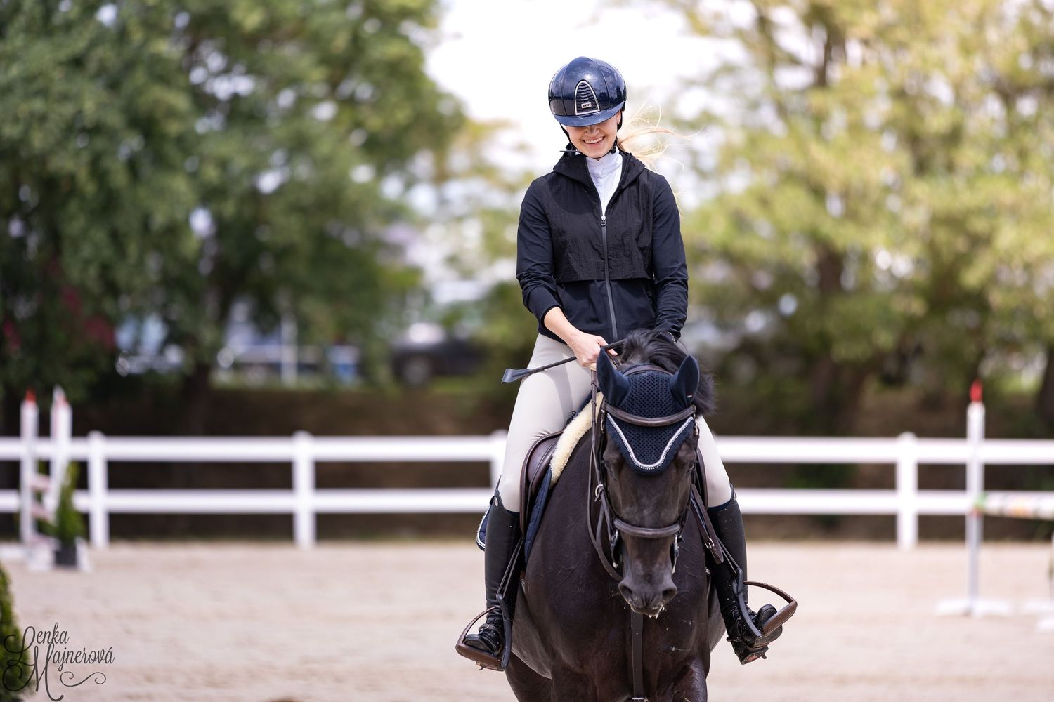 A rider performs dressage movements on a dark horse in an outdoor arena with white fencing.