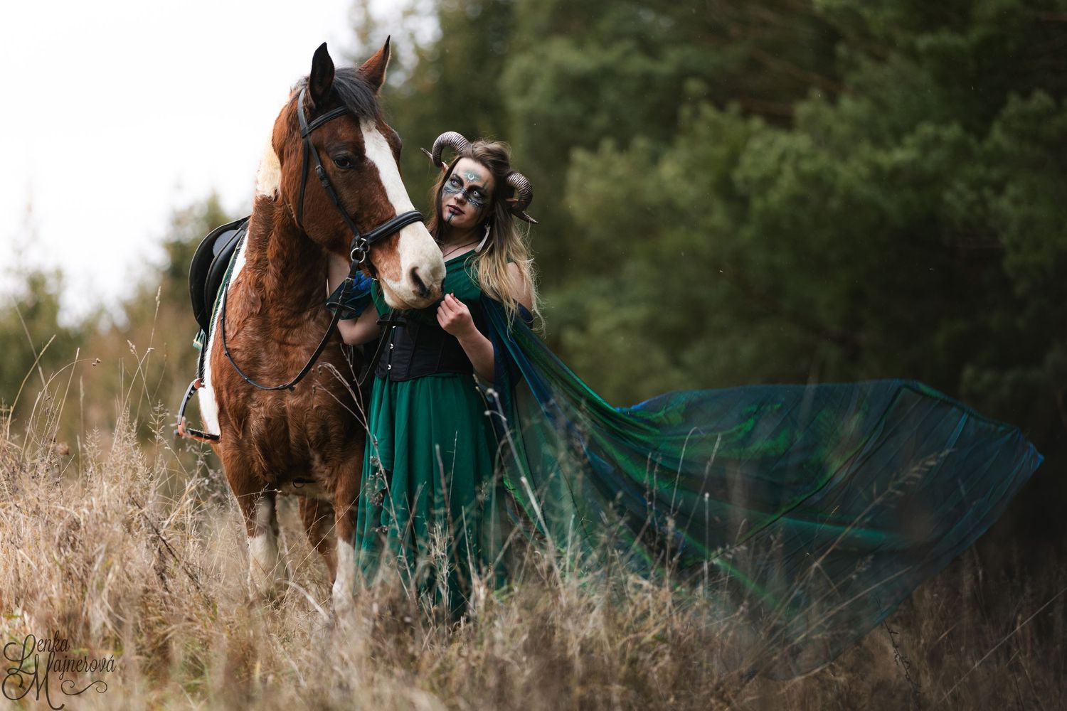 A woman in an emerald green dress walks through a golden field with a brown horse at sunset.
