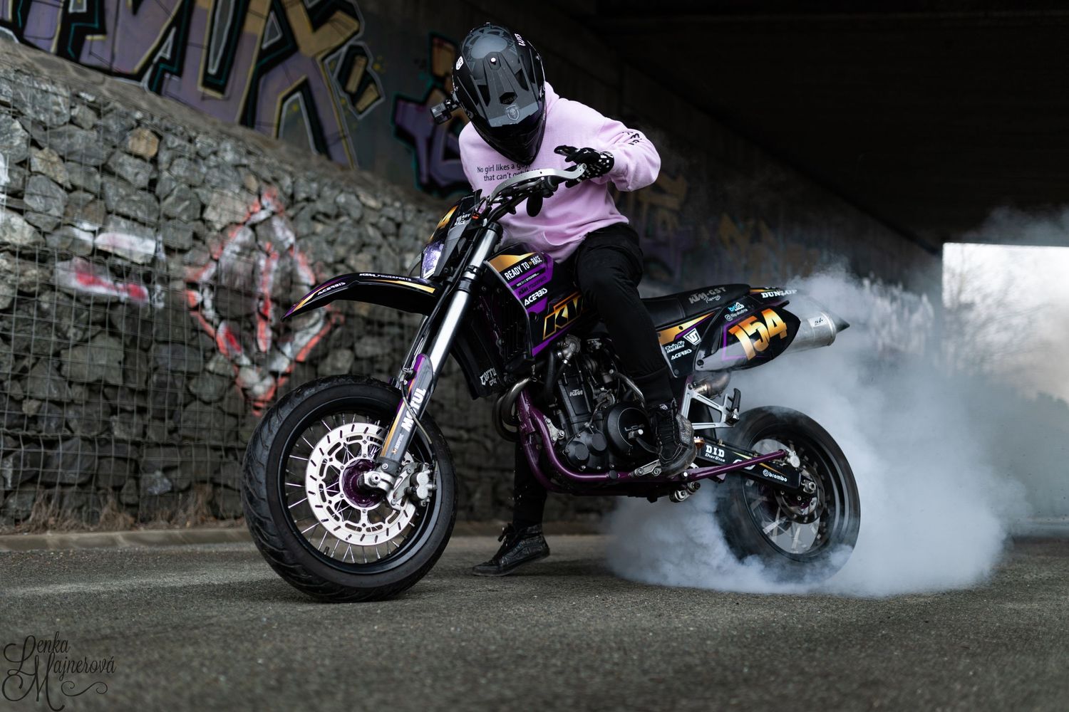 Stunt motorcyclist performing tricks with smoke effects against stone wall at night.
