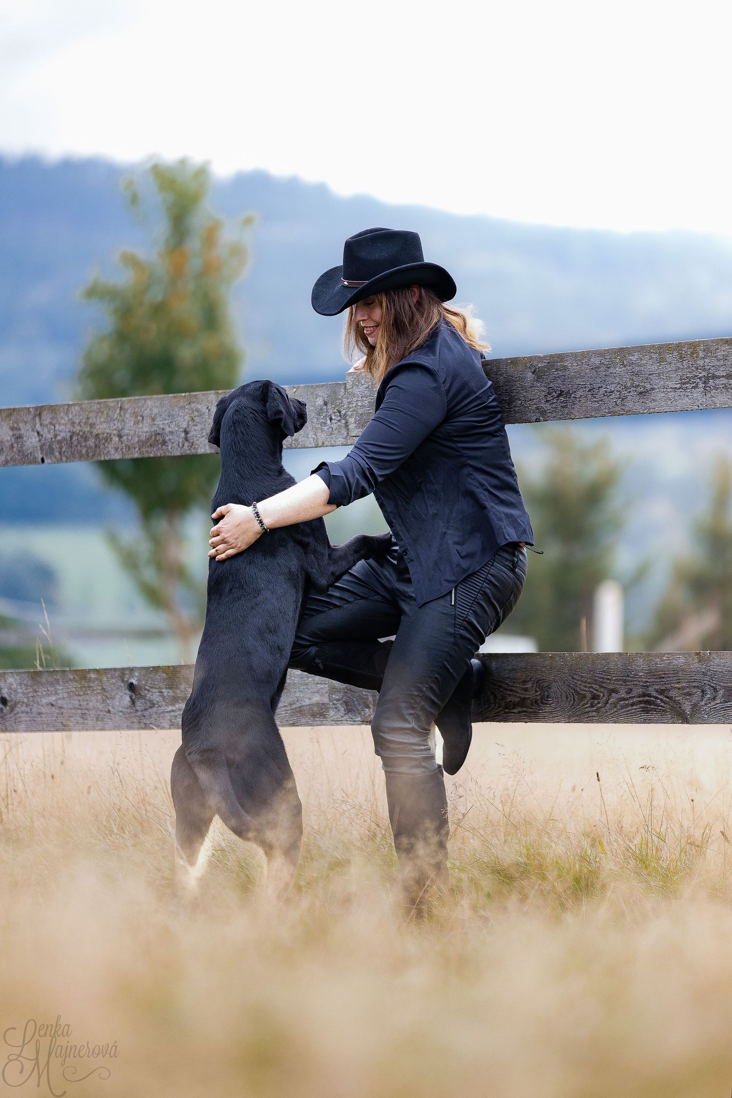 A black dog and person in dark clothing lean against a rustic wooden fence in a rural outdoor setting.