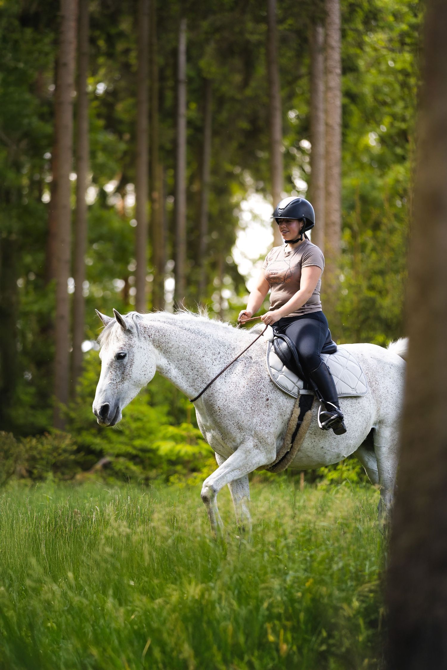 A rider in a helmet and pink shirt rides a dappled gray horse through a sunlit forest trail.
