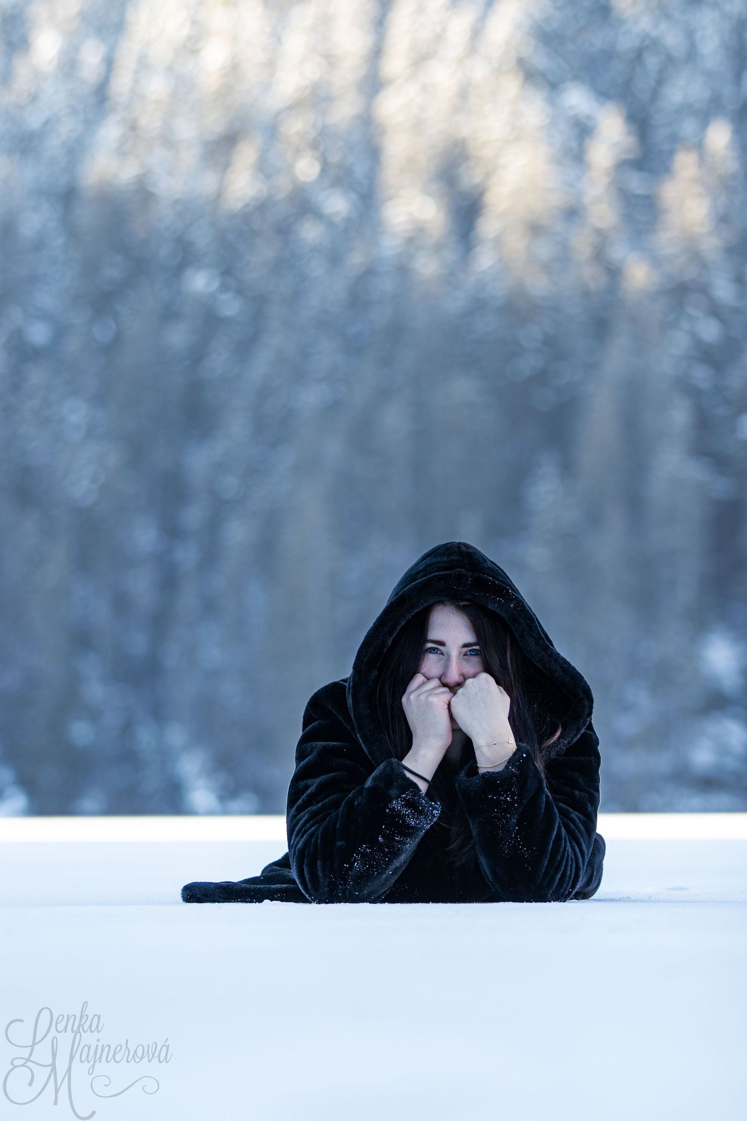 Winter portrait in black hoodie lying on snow with ethereal bokeh background.