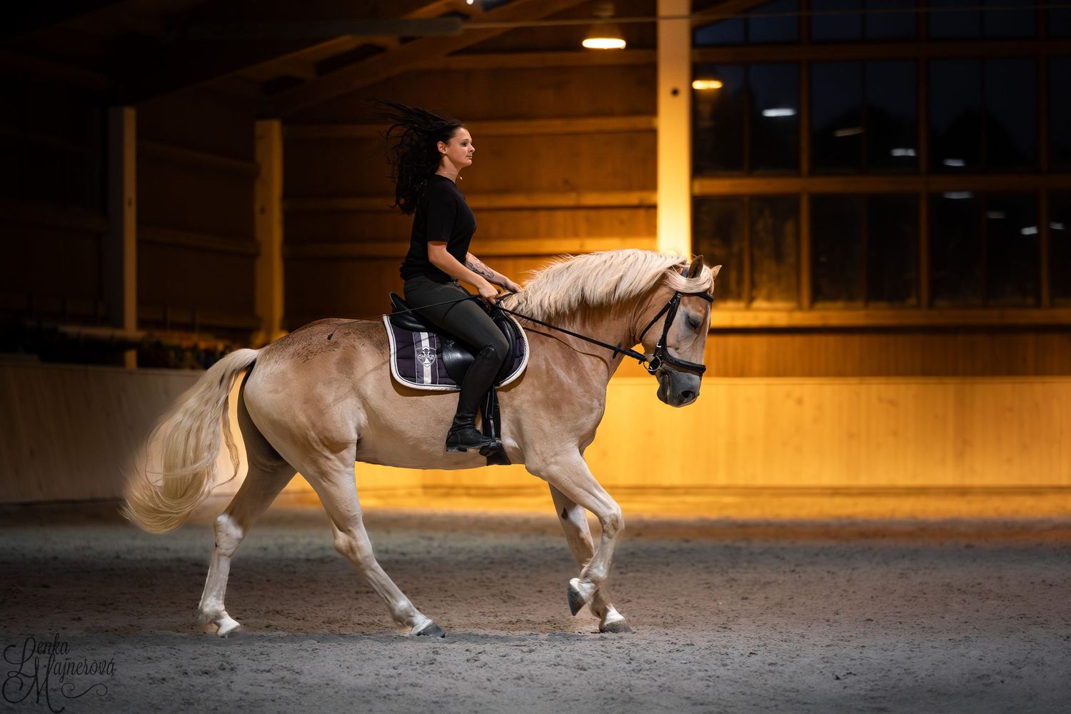 A rider performs a dressage movement on a light-colored horse in an indoor arena with warm lighting.