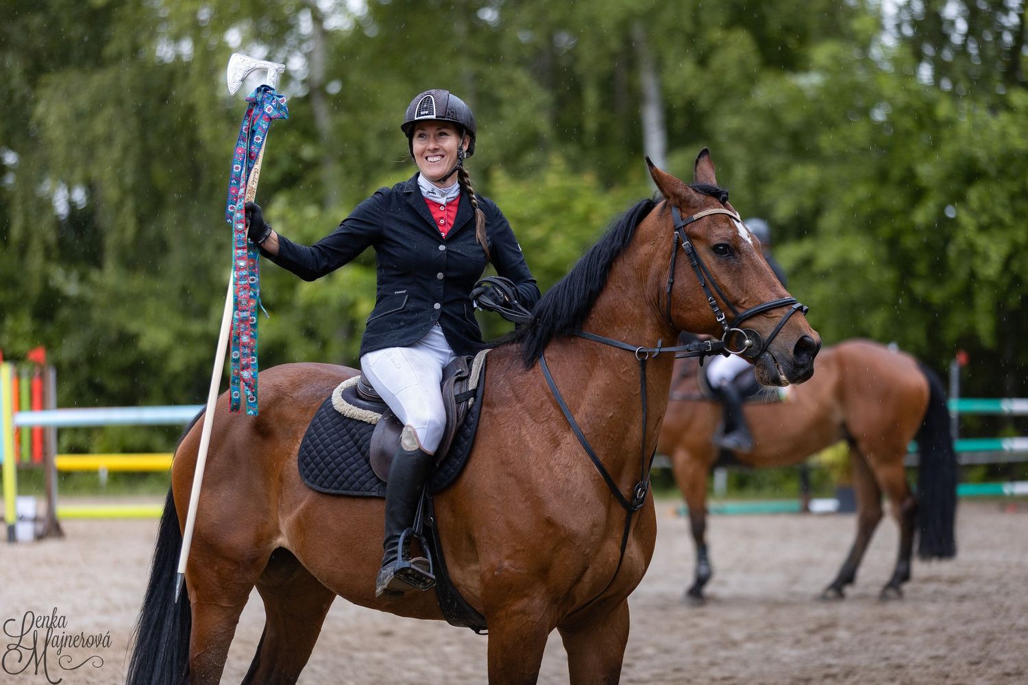 Equestrian riders compete in a jumping competition on horseback at an outdoor arena with green trees in background.