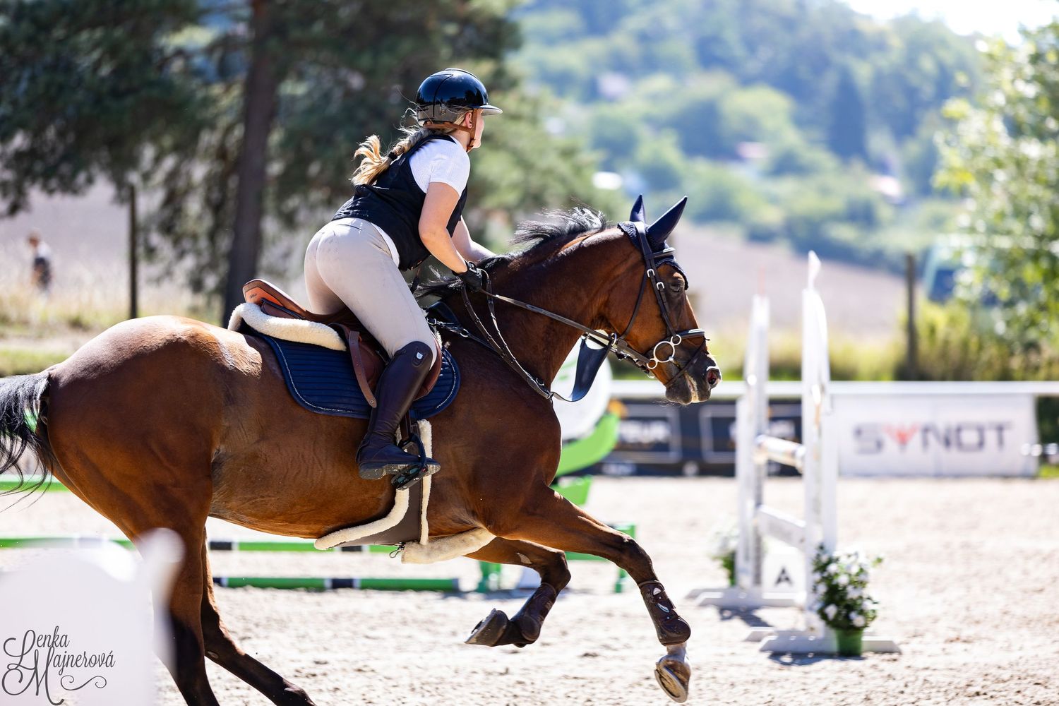 Equestrian rider in navy mask and white attire jumps bay horse over outdoor arena obstacle during competition.