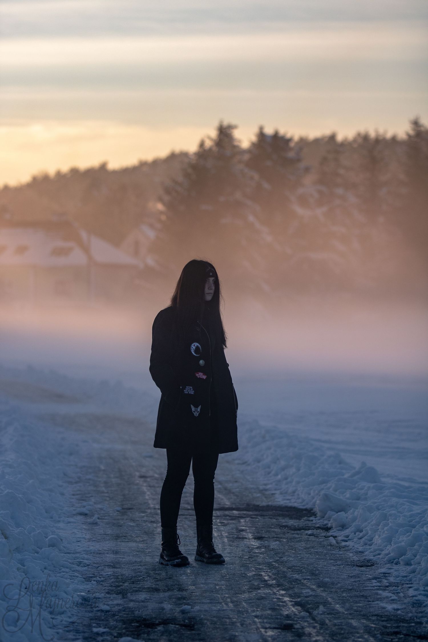 Silhouette of a person standing on a snowy path during a misty sunset with trees in background.